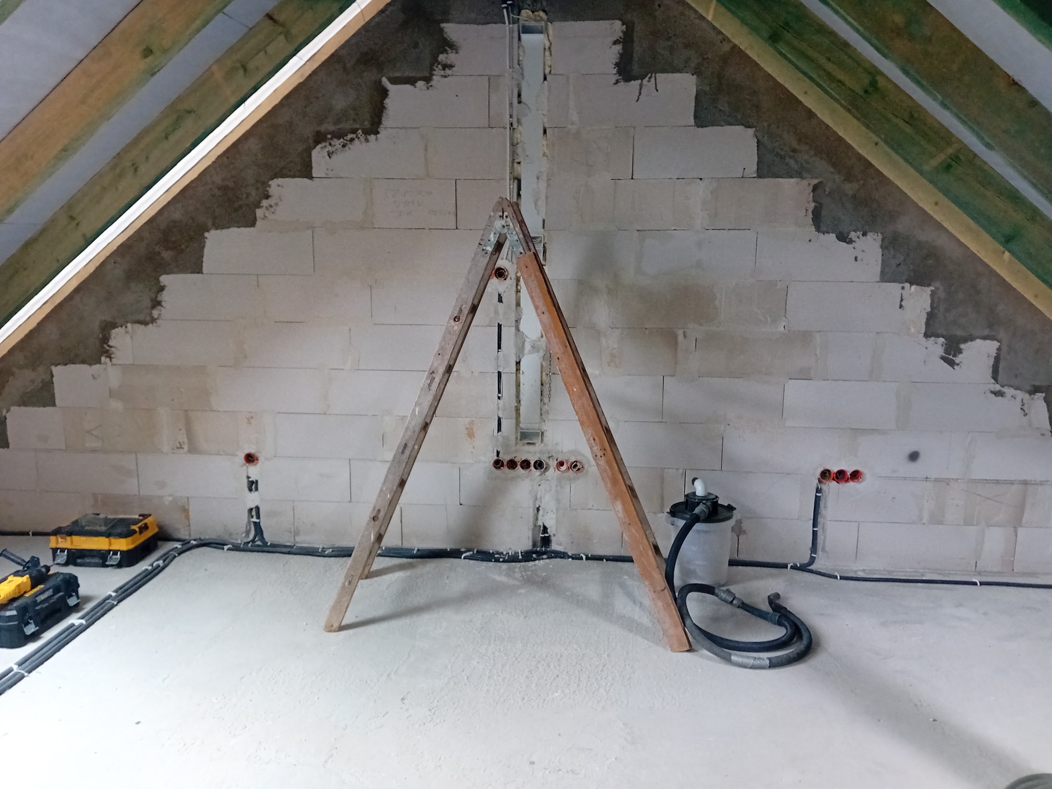 Montaż instalacji elektrycznej w surowym wnętrzu poddasza: widoczne przewody w ścianie z bloczków, puszki elektryczne, drabina i narzędzia na podłodze.