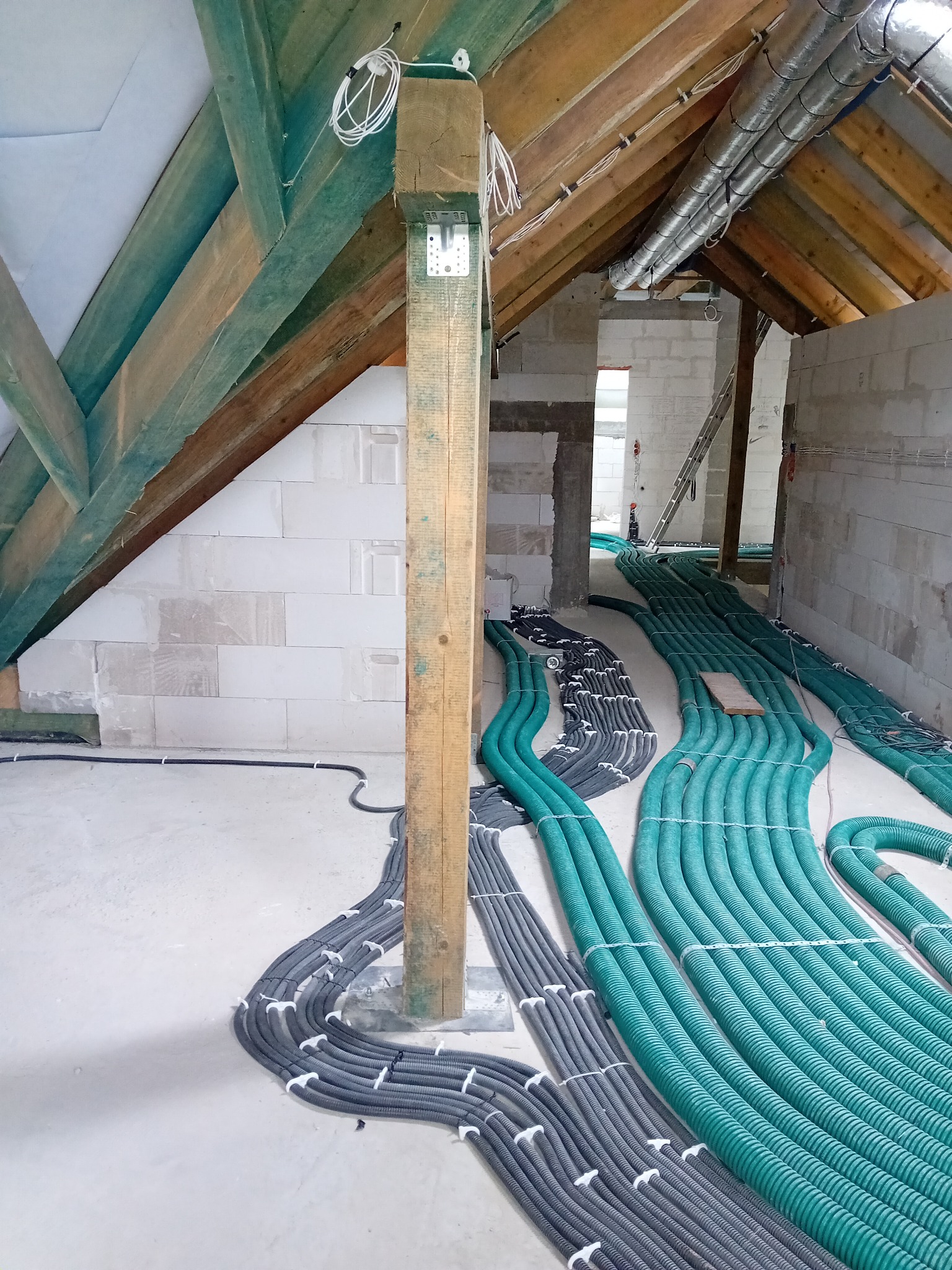 Wnętrze poddasza w trakcie remontu z widocznymi wiązkami kabli elektrycznych w peszlach, częściowo w kolorze zielonym i szarym, ułożonych równolegle na podłodze wzdłuż ściany z białych bloczków,...