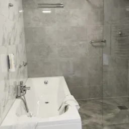 Nowoczesna łazienka z białą wanną, szklaną kabiną prysznicową i szarymi płytkami imitującymi kamień, akcent marmurowy na ścianie, chromowana armatura i akcesoria.