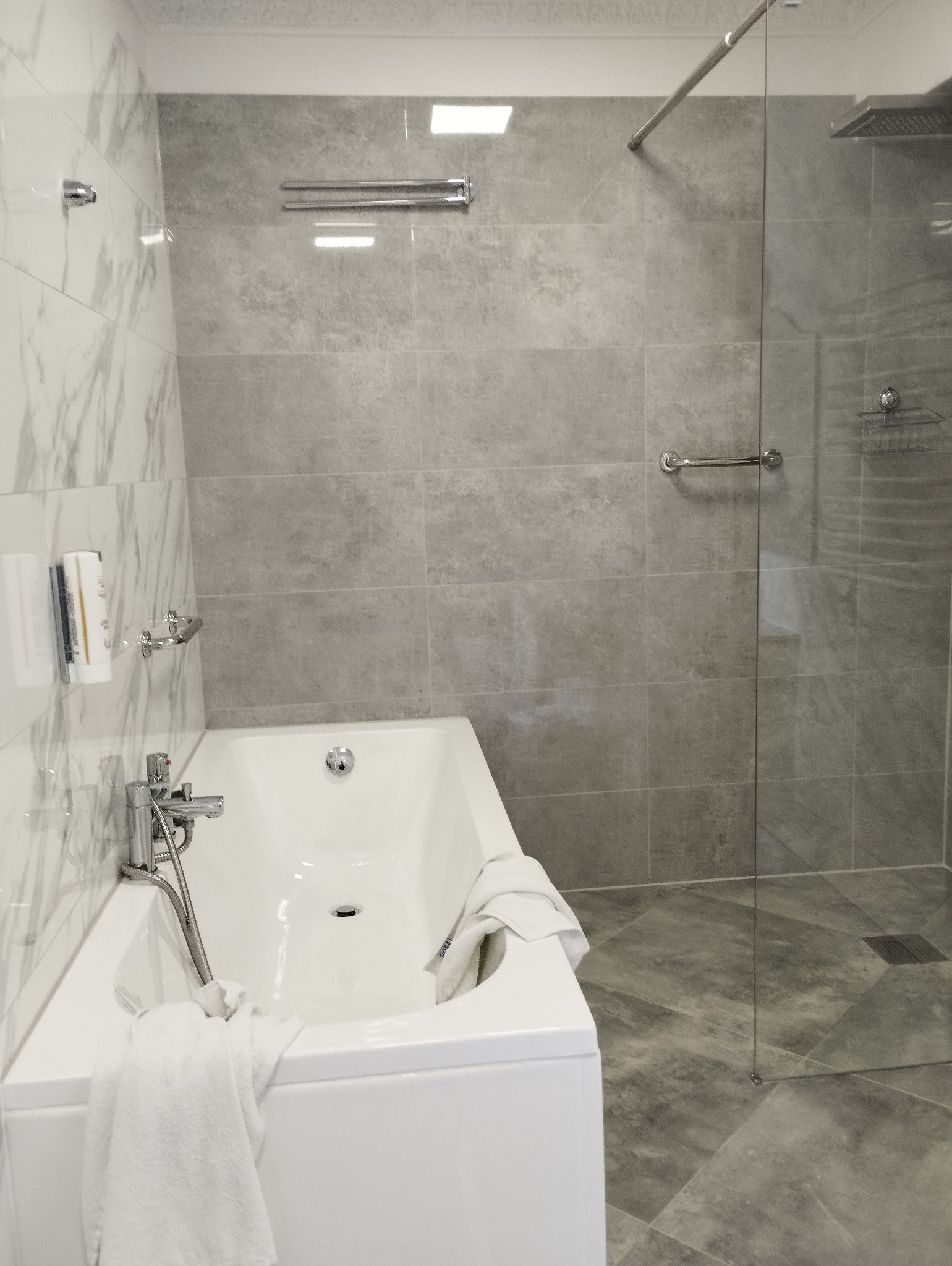 Nowoczesna łazienka z białą wanną, szklaną kabiną prysznicową i szarymi płytkami imitującymi kamień, akcent marmurowy na ścianie, chromowana armatura i akcesoria.