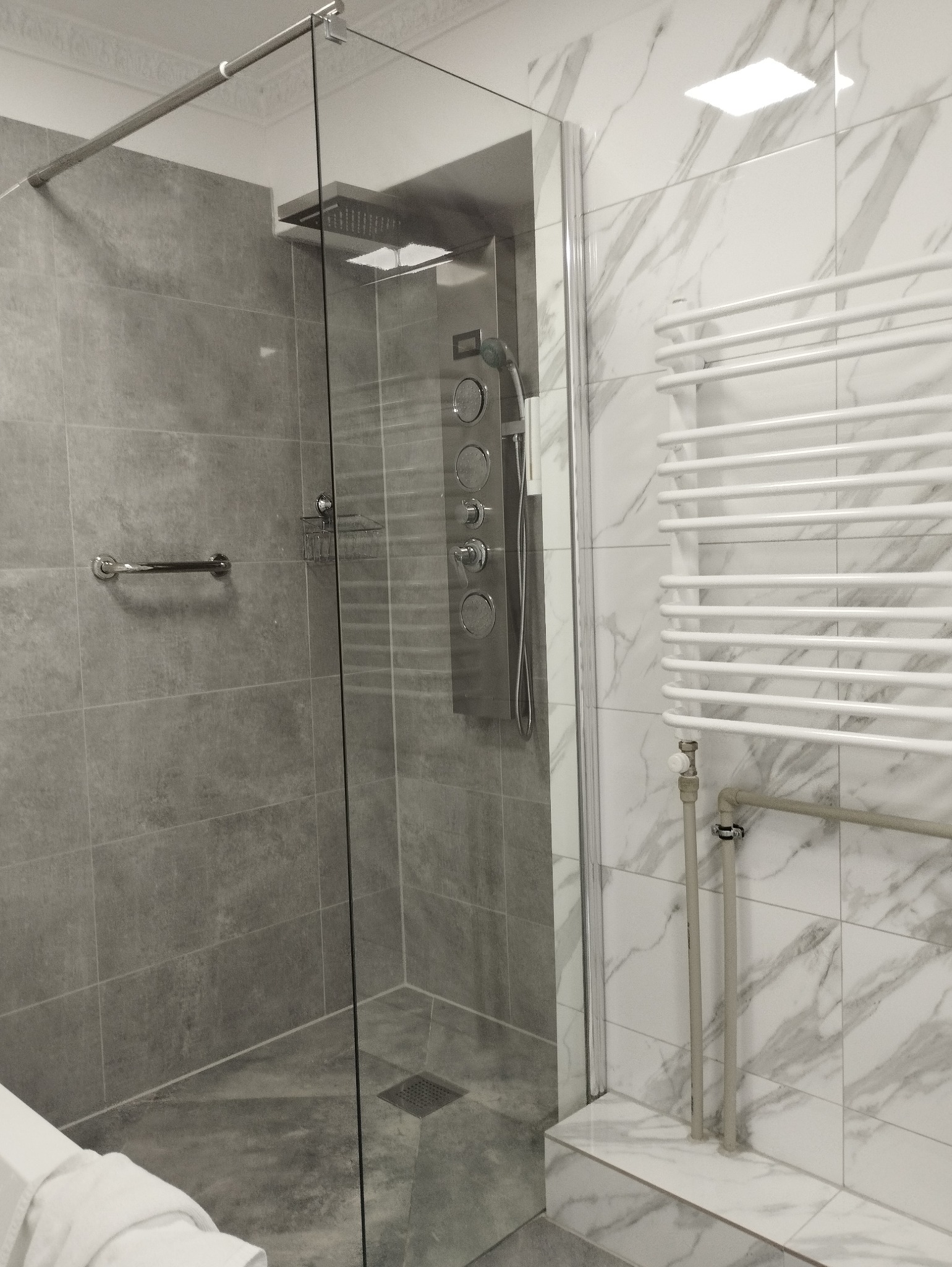 Nowoczesna kabina prysznicowa typu walk-in z szarymi płytkami imitującymi beton i białymi płytkami imitującymi marmur, wyposażona w panel prysznicowy z hydromasażem i grzejnik drabinkowy.