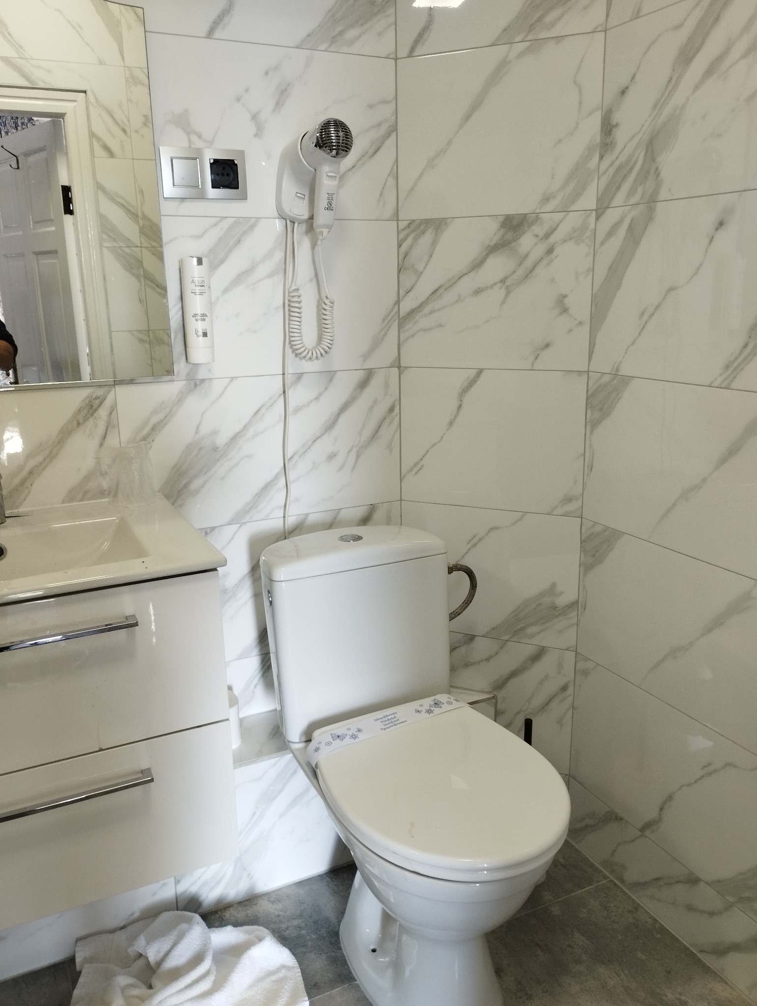 Nowoczesna łazienka z białymi marmurkowymi płytkami, wiszącą suszarką do włosów, toaletą z higieniczną nakładką na deskę i umywalką z szafką, widoczna w lustrze część pokoju.