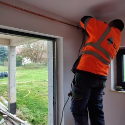 Instalacje Gazowe KACPER WOJTEK - Instalator w pomarańczowej kamizelce montuje miedzianą rurę pod sufitem w surowym wnętrzu, widok przez okno na zewnątrz.