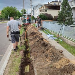 Instalacje Gazowe KACPER WOJTEK - Prace ziemne: wykop pod pomarańczową rurę, widoczni dwaj pracownicy i minikoparka na tle ulicy i budynków.