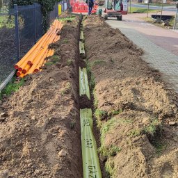 Instalacje Gazowe KACPER WOJTEK - Wykop z ułożonym wewnątrz rurociągiem gazowym w zielonej folii ochronnej z napisem 'GAZ', obok pomarańczowe rury, w tle pracownik i minikoparka.