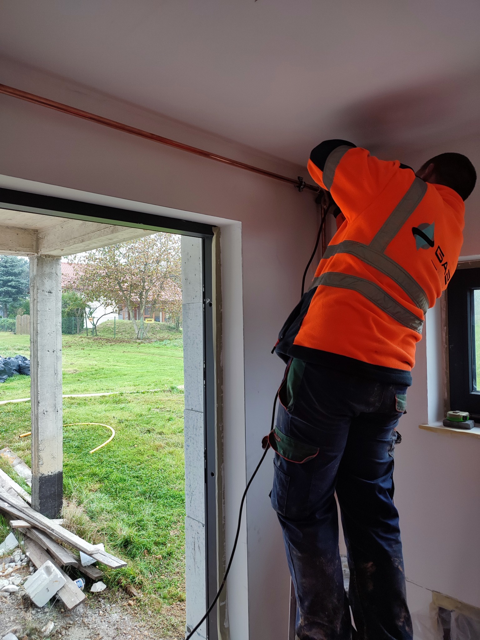 Instalator w pomarańczowej kamizelce montuje miedzianą rurę pod sufitem w surowym wnętrzu, widok przez okno na zewnątrz.