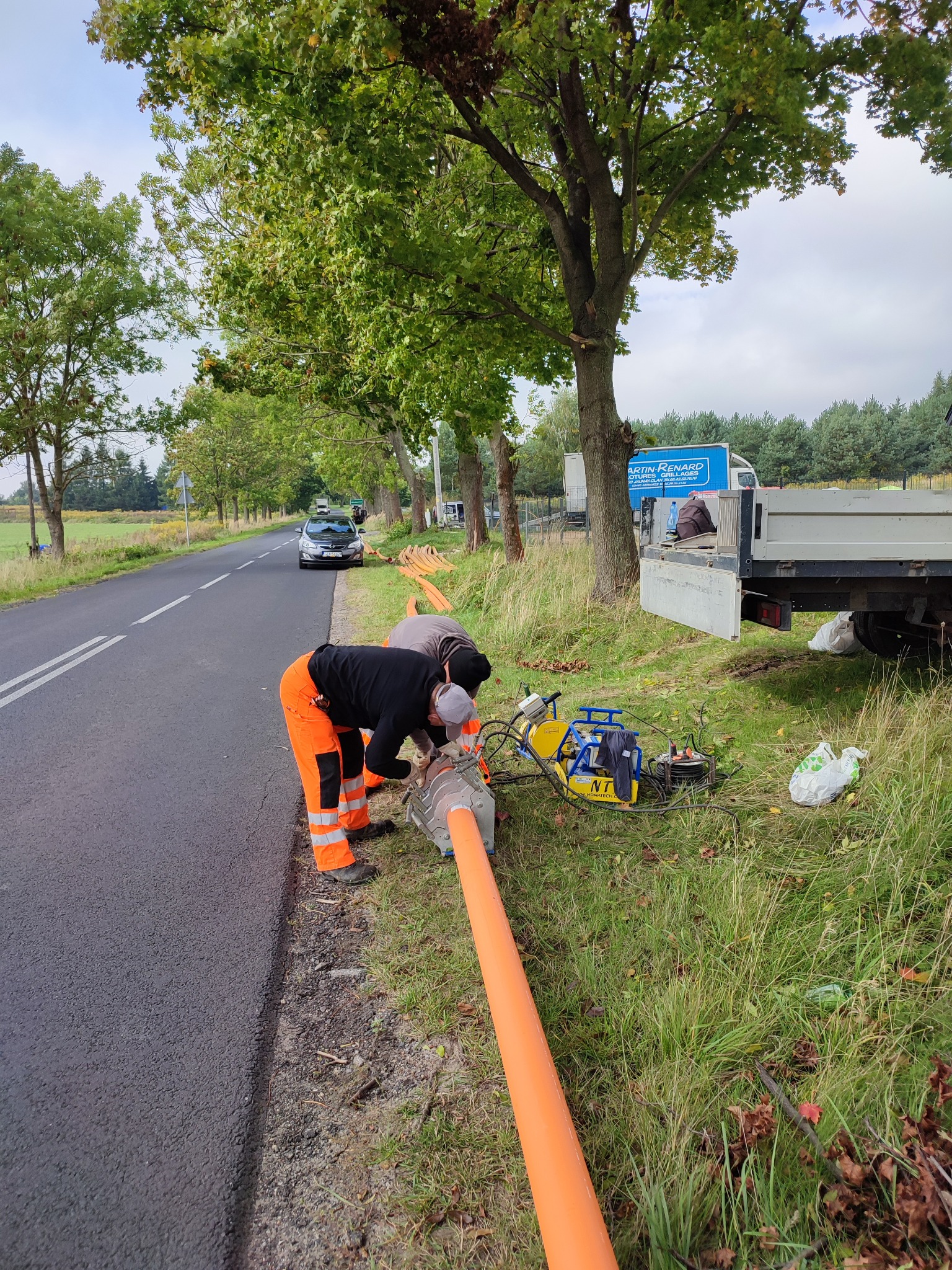 Dwóch pracowników w pomarańczowych kombinezonach łączy pomarańczową rurę na poboczu drogi w Skoczowie, w tle widoczny samochód i ciężarówka z plandeką.