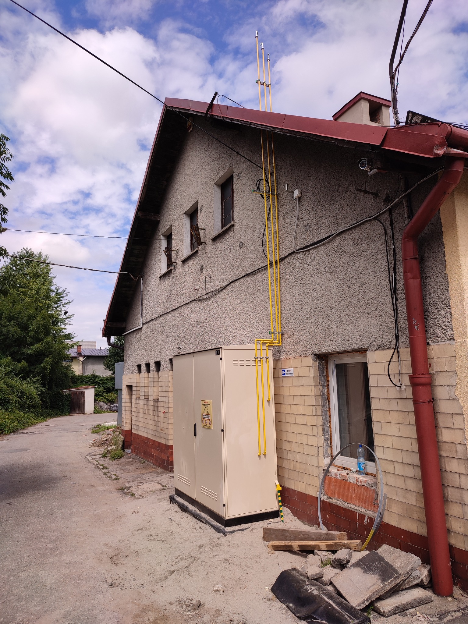 Elewacja budynku z nową instalacją gazową, rury w kolorze żółtym prowadzone wzdłuż ściany i do góry, widoczna szafka gazowa przy budynku, fragment chodnika i nieba z chmurami.