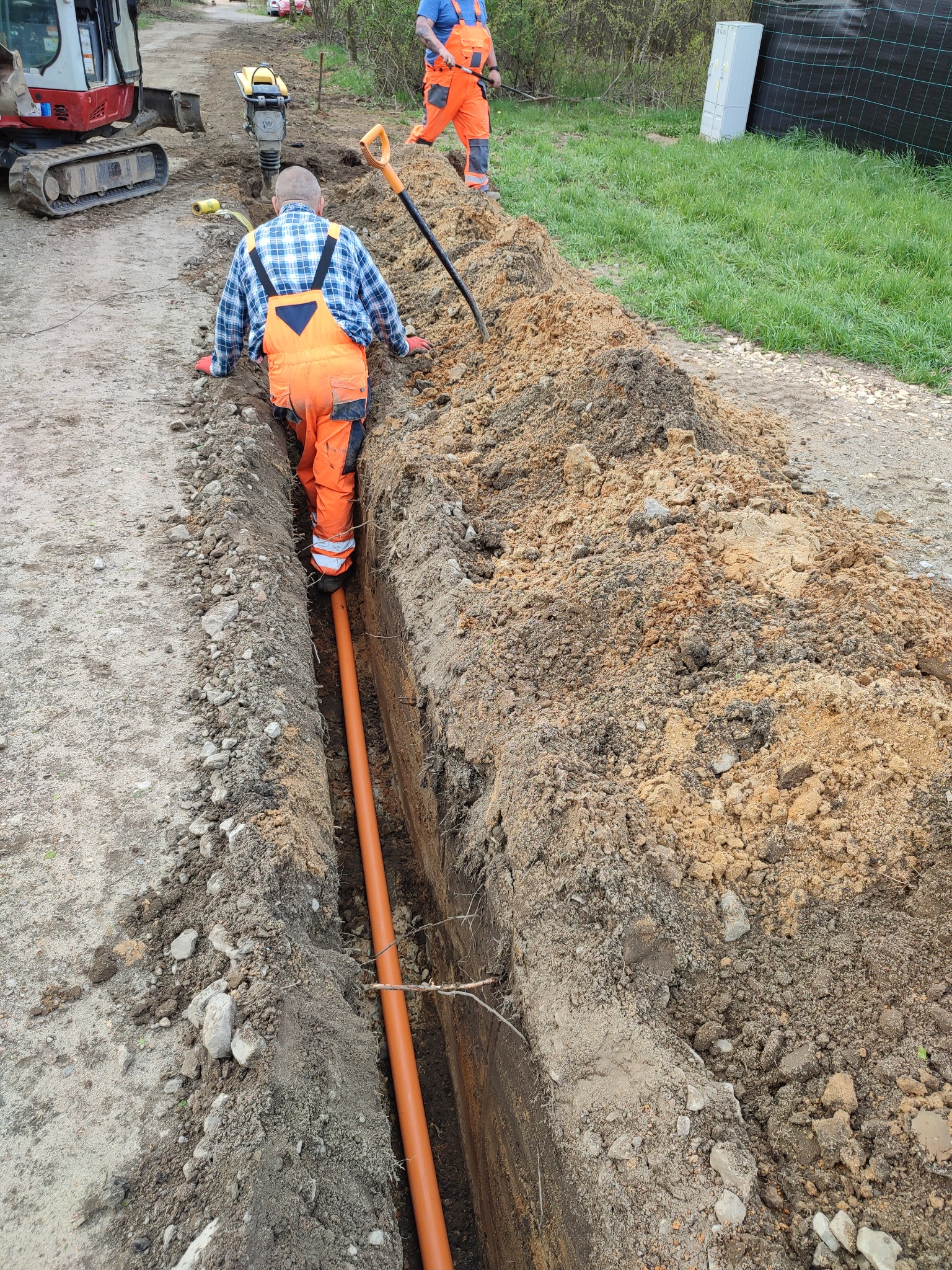 Montaż pomarańczowej rury w wykopie, widoczni dwaj pracownicy w pomarańczowych kombinezonach, w tle mini koparka i ubijarka gruntu.