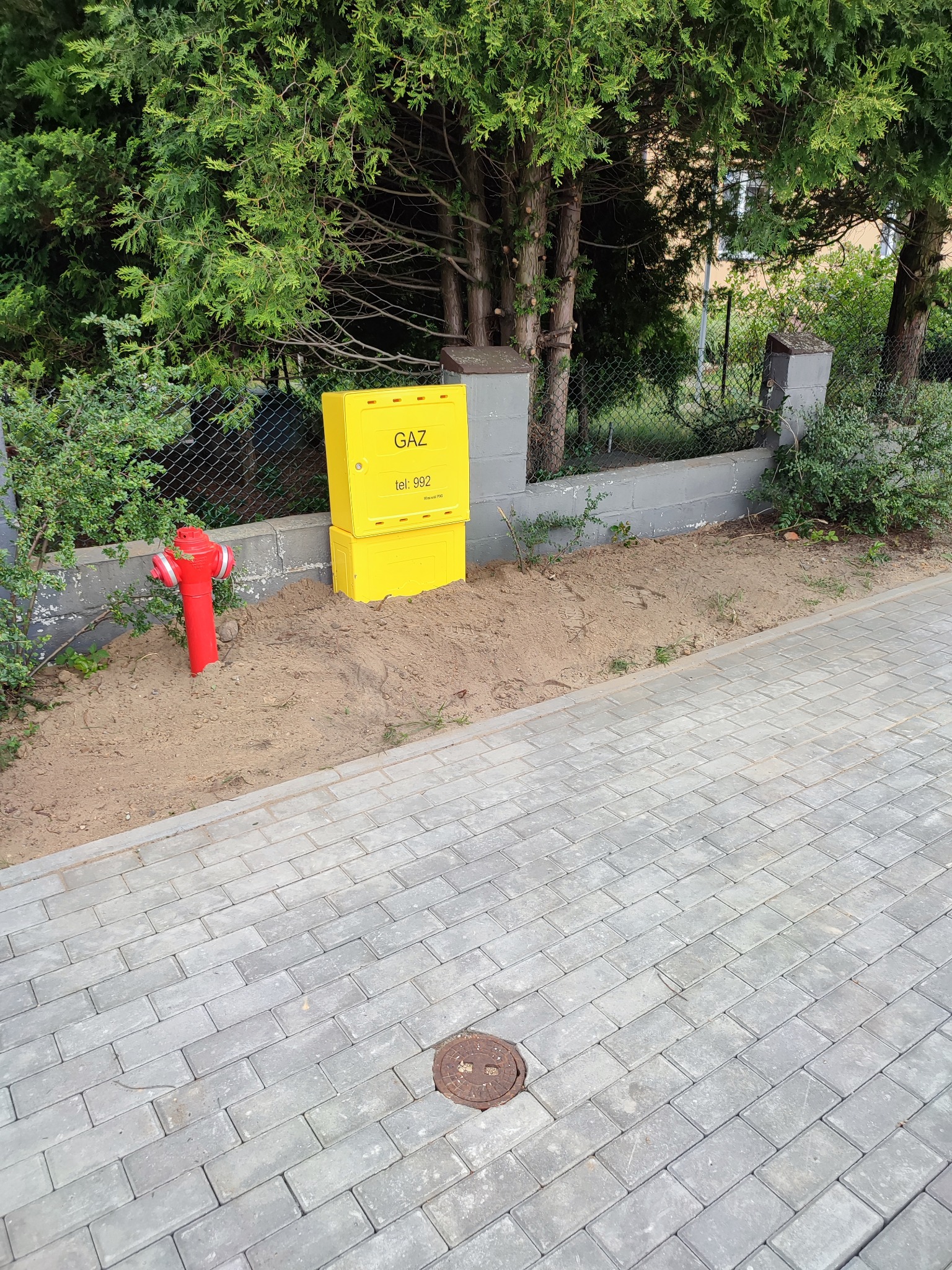 Żółta skrzynka gazowa z numerem telefonu, umieszczona na trawniku obok czerwonego hydrantu, widok z poziomu chodnika z szarej kostki.