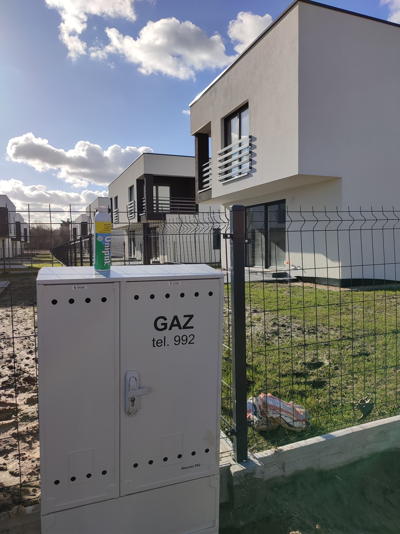 Biała szafka gazowa z napisem 'GAZ tel. 992' na tle nowoczesnych domów jednorodzinnych i ogrodzenia, z butelką środka uszczelniającego Unipak na górze.