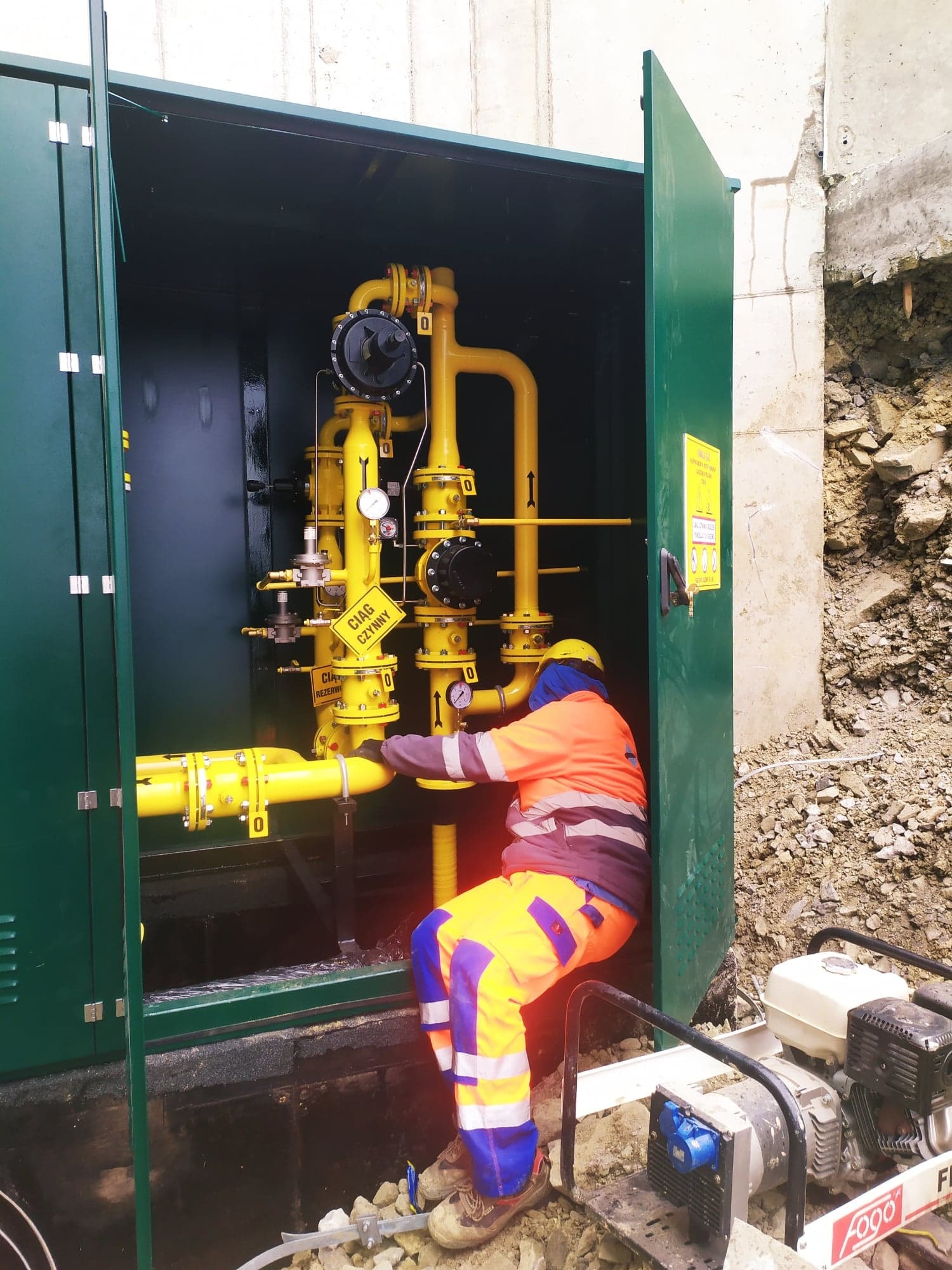 Pracownik w odzieży roboczej, wewnątrz zielonej szafki z żółtymi rurami gazowymi, podłączający instalację; widoczne oznaczenia 'Ciąg czynny' i agregat prądotwórczy na zewnątrz.