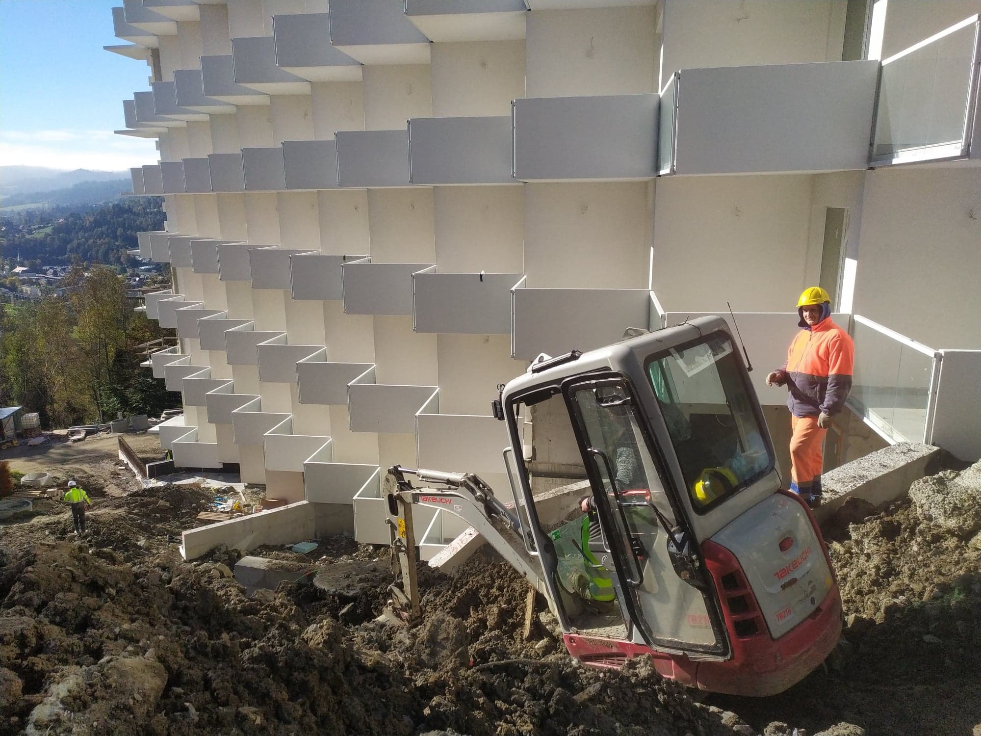 Mini koparka Takeuchi TB216 podczas prac ziemnych przy budynku z balkonami w kształcie kostek. Pracownik w odblaskowej odzieży stoi obok koparki. W tle widoczne drzewa i zabudowa miejska.