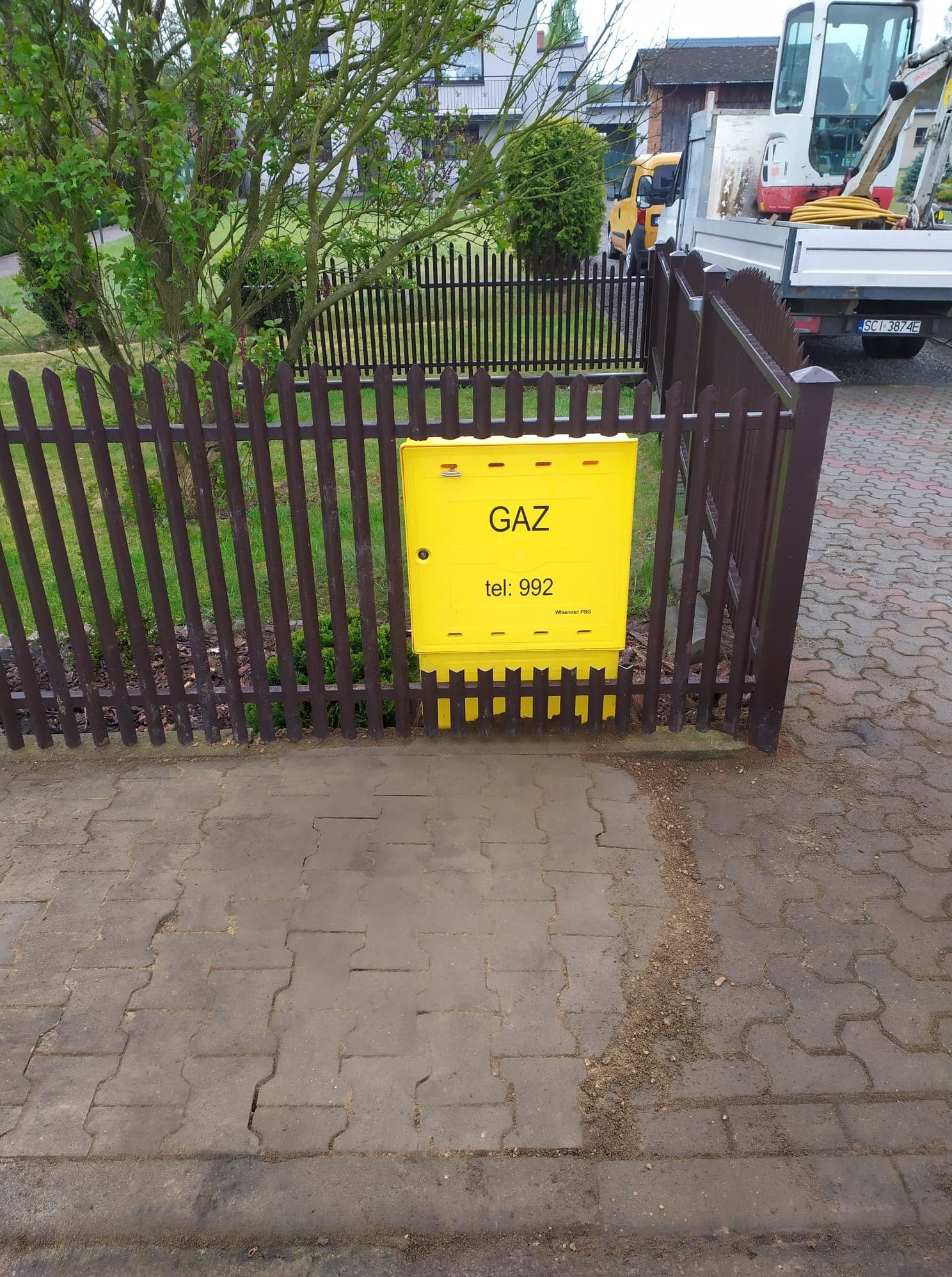 Żółta skrzynka gazowa z napisem 'GAZ' i numerem telefonu, zamontowana na brązowym ogrodzeniu z metalowych sztachet, na tle zieleni i częściowo widocznego pojazdu budowlanego.