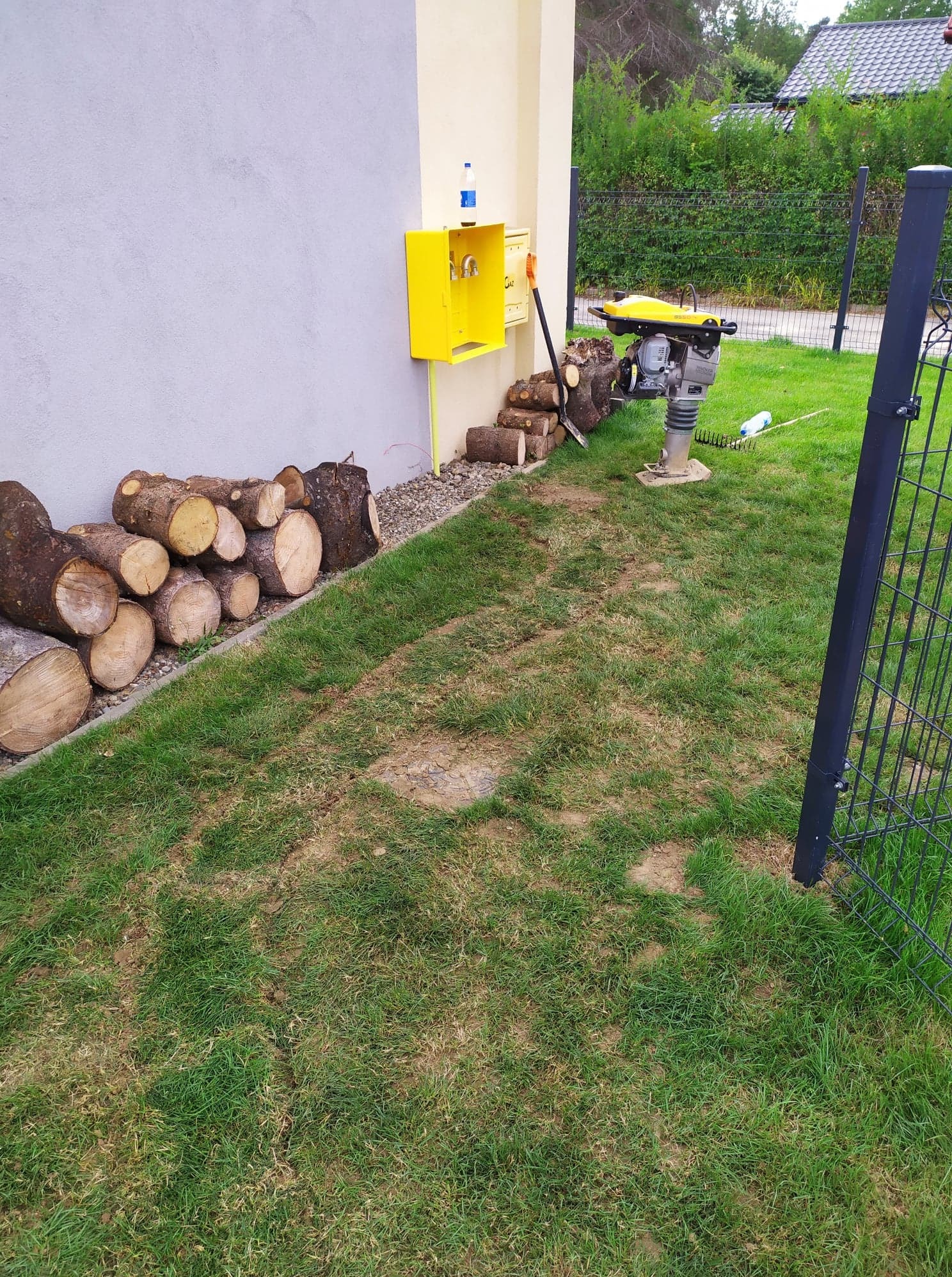 Żółta skrzynka gazowa na ścianie budynku, obok stos drewna, ubijarka wibracyjna na trawniku i fragment ogrodzenia.