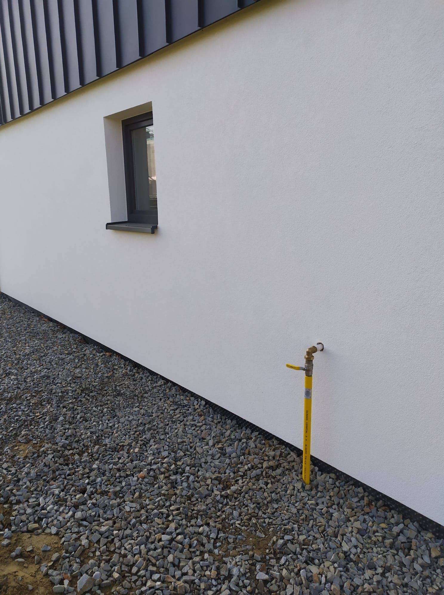 Żółty zawór gazowy wystający ze ściany budynku, widoczne kamienie przy fundamencie, małe okno w ścianie.