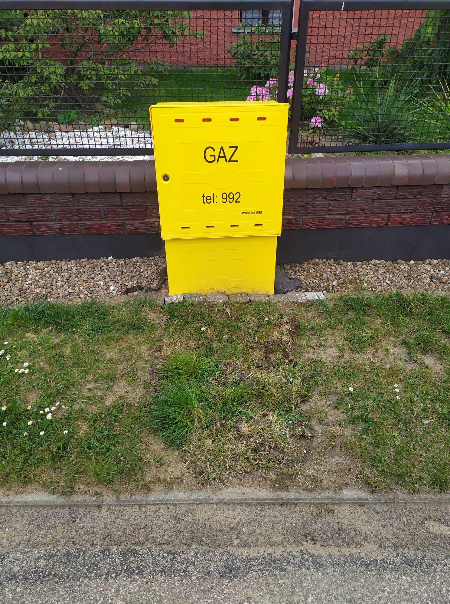 Żółta skrzynka gazowa z napisem 'GAZ' i numerem telefonu, umieszczona na tle ceglanego murku, siatki ogrodzeniowej i zieleni