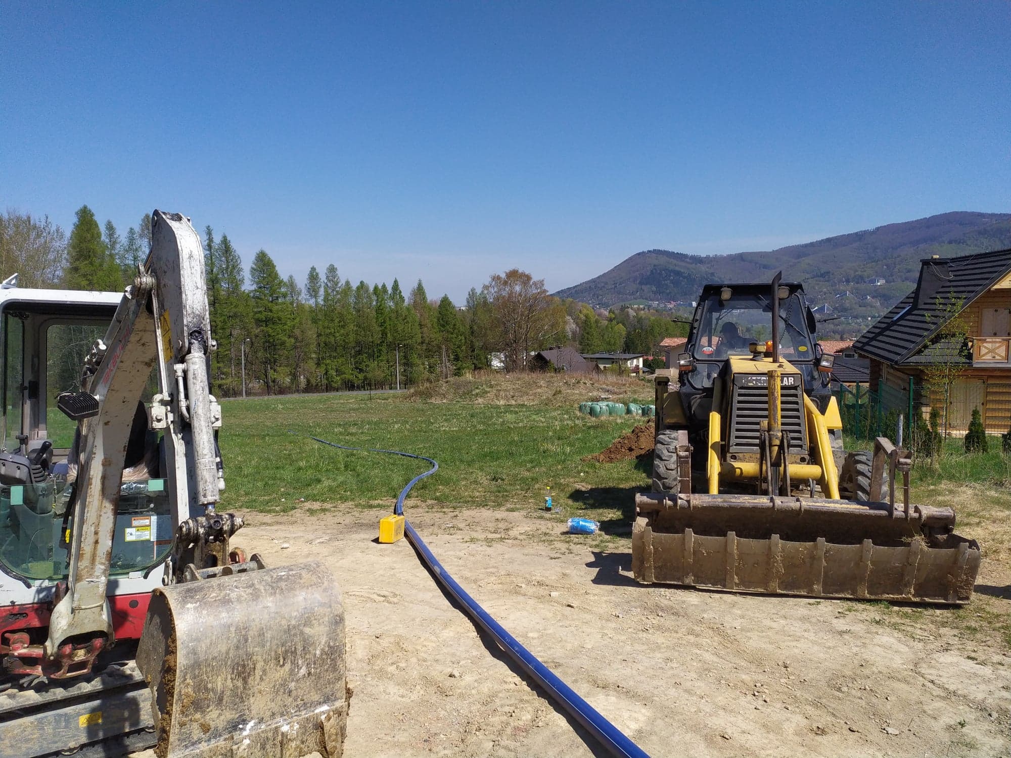 Dwa ciężkie pojazdy budowlane, koparka i ładowarka, przygotowują teren pod instalację rury w malowniczym krajobrazie z domem i górami w tle.