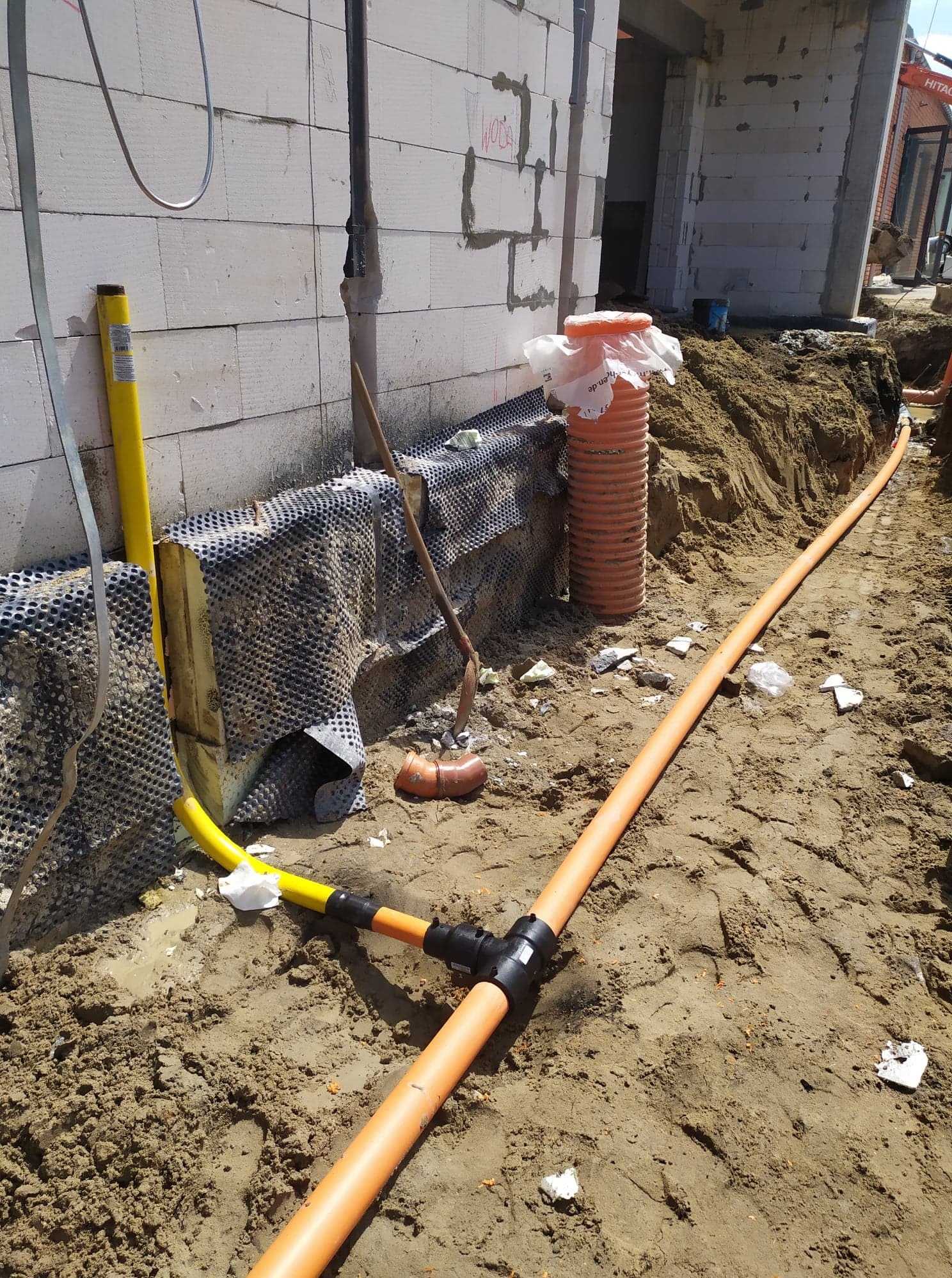 Pomarańczowe rury gazowe z czarnymi łącznikami w wykopie przy fundamencie budynku z białego betonu komórkowego, widoczna folia kubełkowa i żółta rura osłonowa.