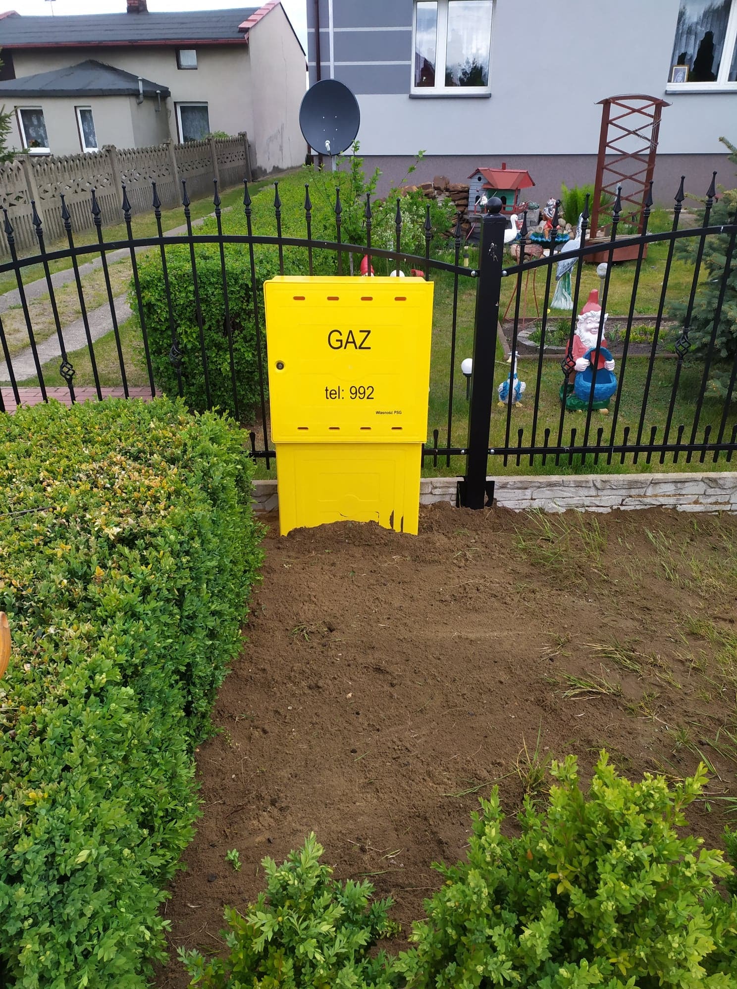 Żółta skrzynka gazowa z numerem telefonu na tle czarnego ogrodzenia i zadbanego ogrodu.
