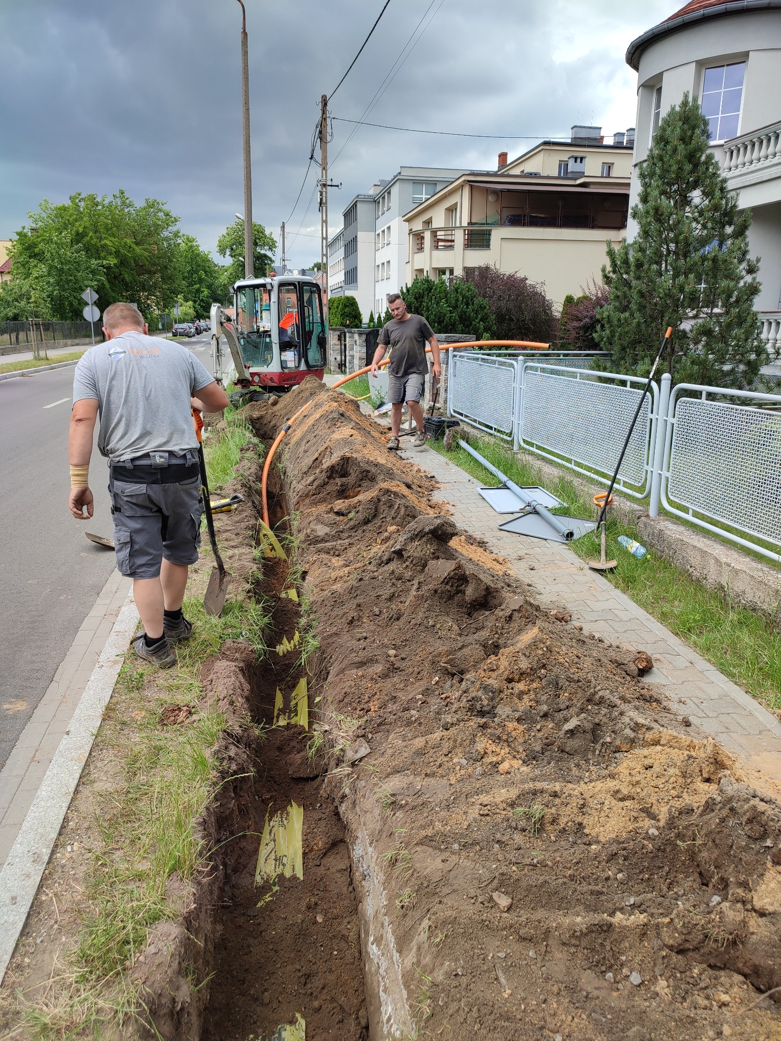 Prace ziemne: wykop pod pomarańczową rurę, widoczni dwaj pracownicy i minikoparka na tle ulicy i budynków.