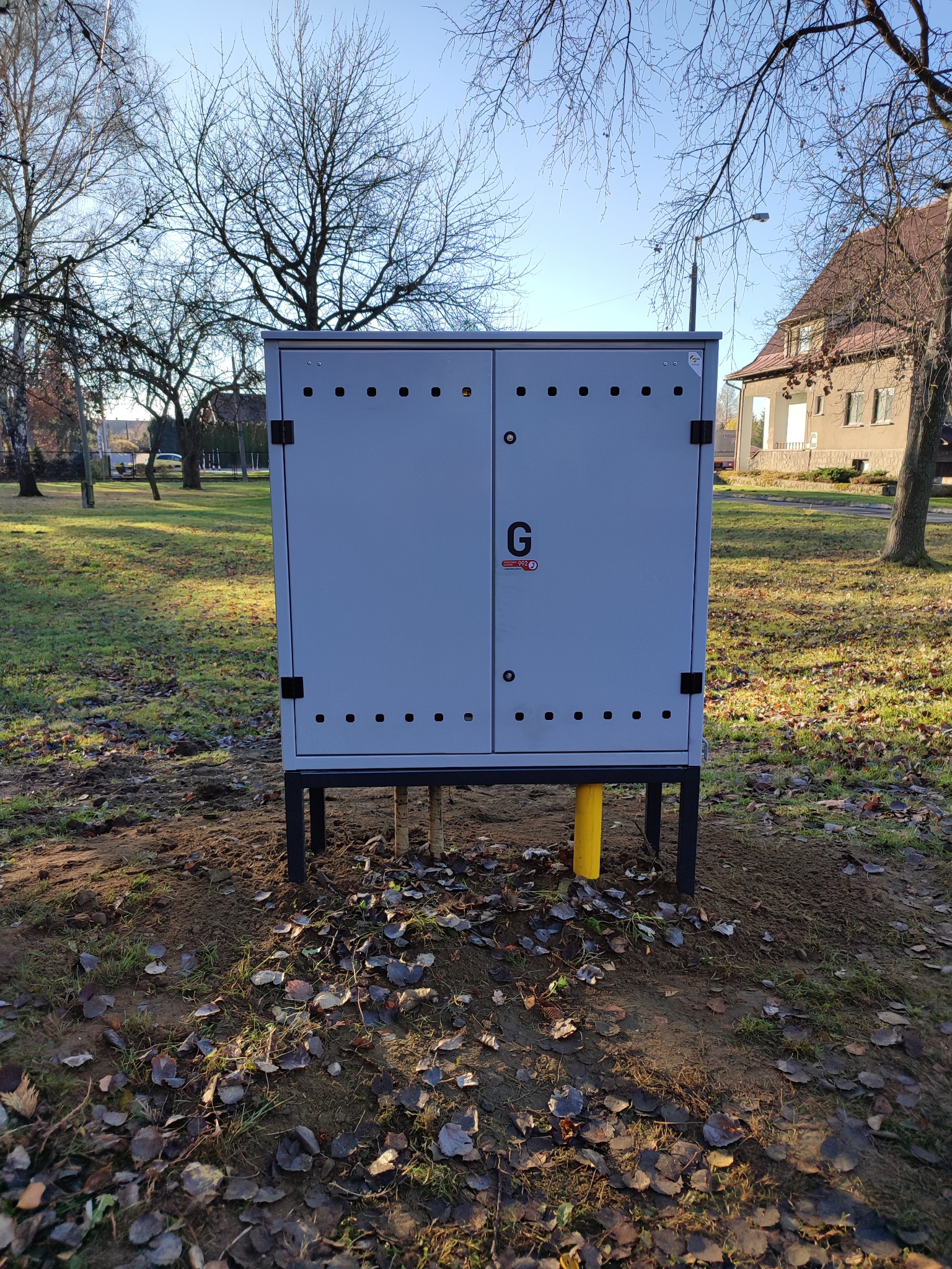 Szara szafka gazowa na czarnych nóżkach z żółtym wystającym rurociągiem na zewnątrz budynku, na trawniku pokrytym liśćmi.