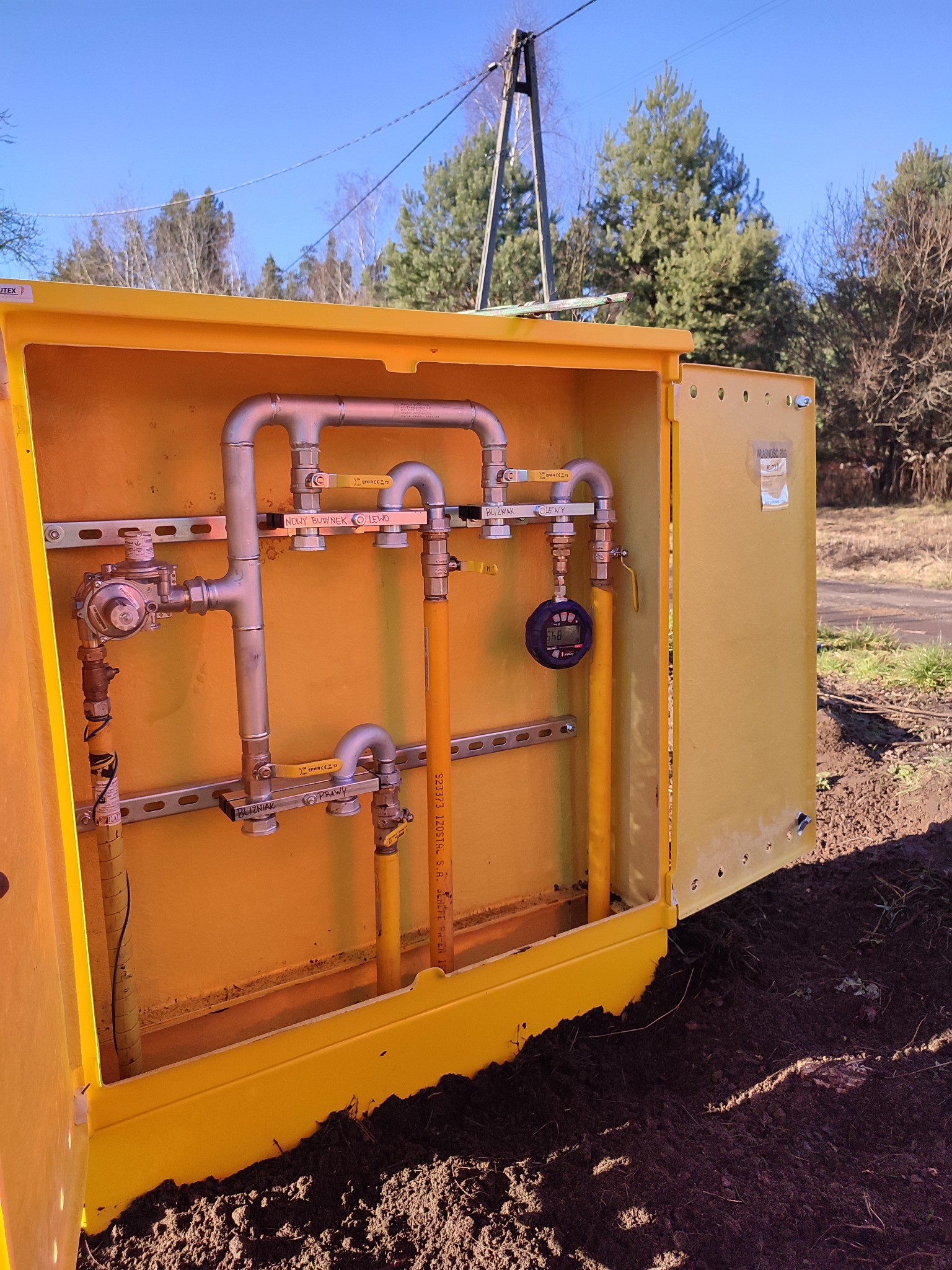 Żółta szafka gazowa z otwartymi drzwiami, widoczne wewnętrzne rury gazowe, zawory z opisami 'Nowy budynek lewo', 'Bliźniak prawy', 'Bliźniak lewy' i manometr cyfrowy. Szafka osadzona w ziemi...