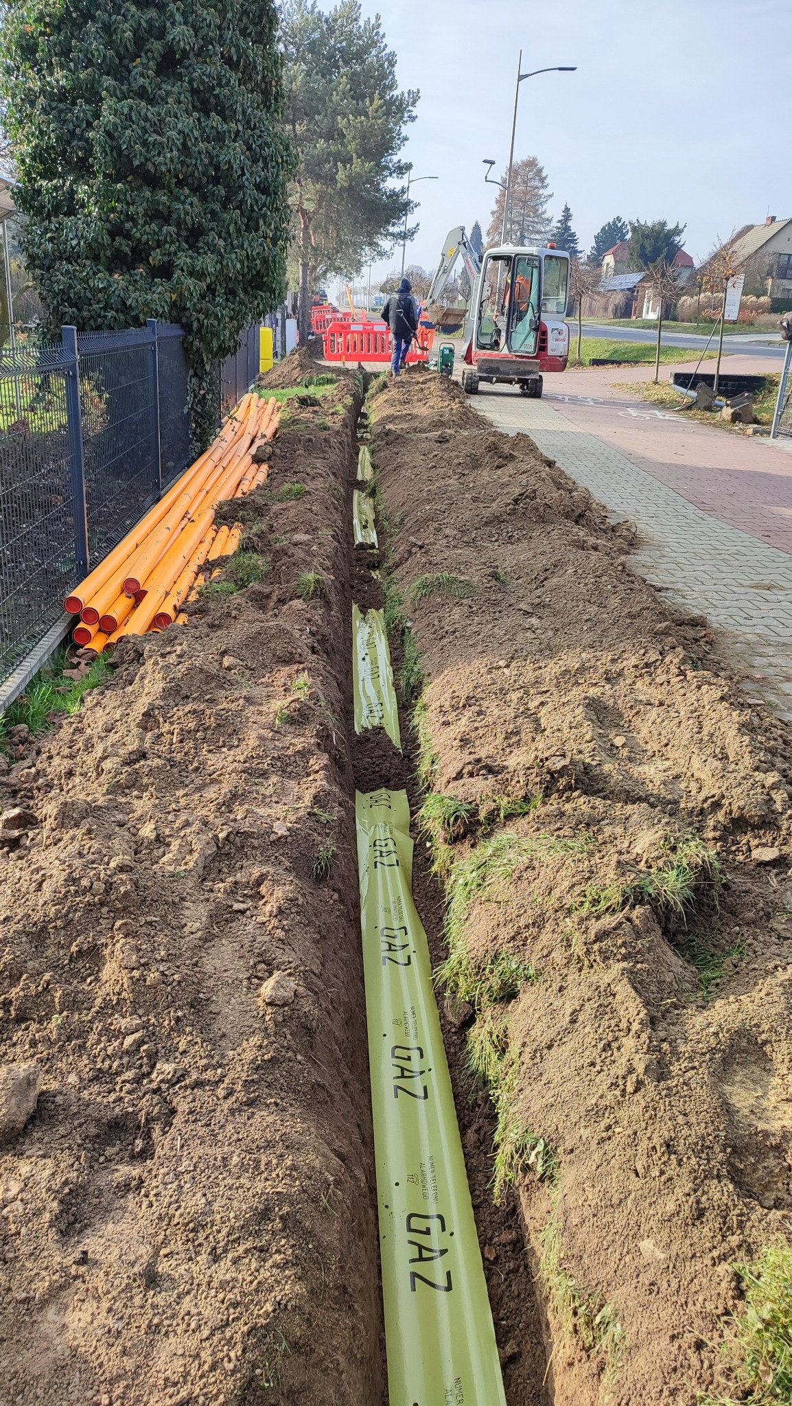 Wykop z ułożonym wewnątrz rurociągiem gazowym w zielonej folii ochronnej z napisem 'GAZ', obok pomarańczowe rury, w tle pracownik i minikoparka.