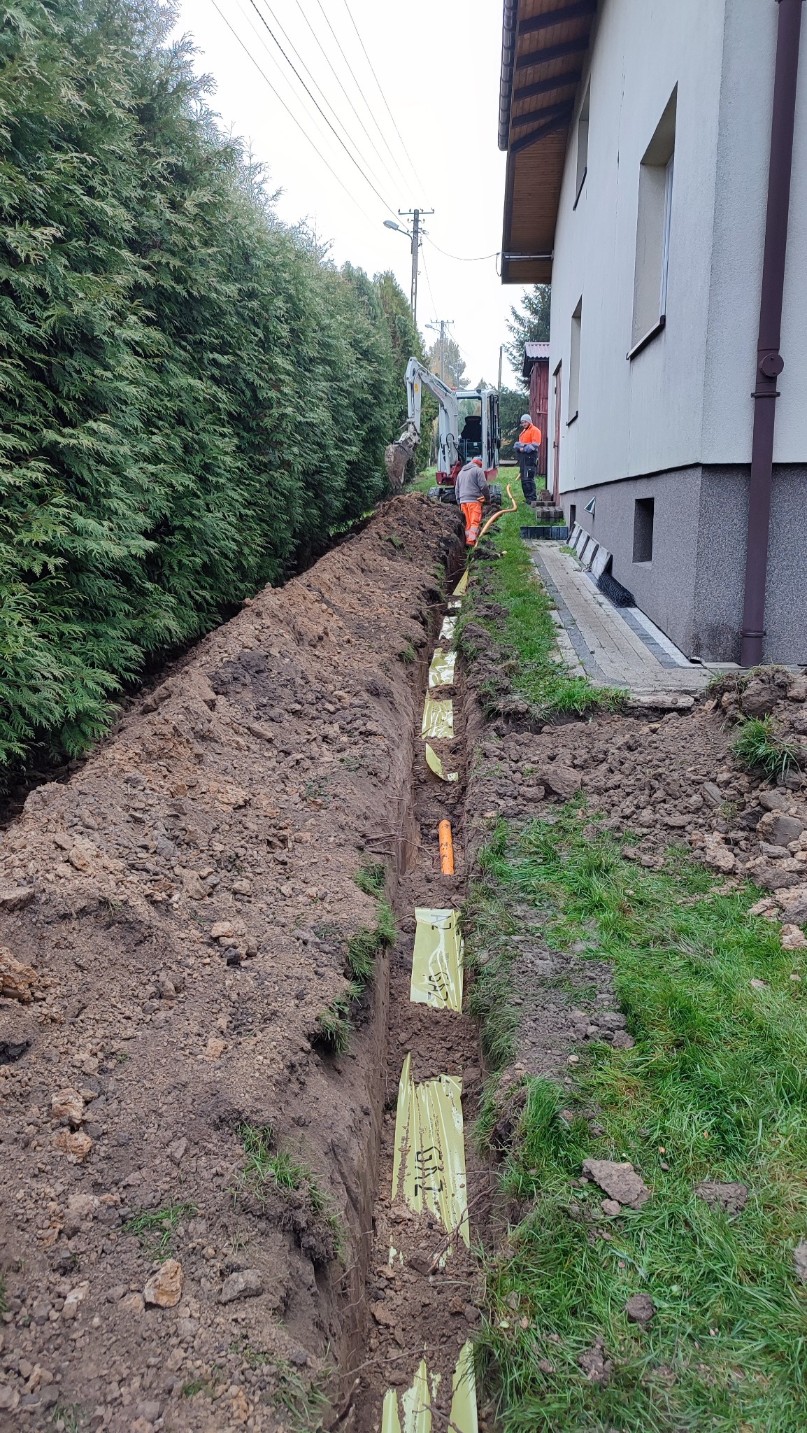Wykop pod instalację, widoczne pomarańczowe rury i żółta folia ostrzegawcza, w tle pracujący mały koparka i dwóch robotników w pomarańczowych ubraniach.