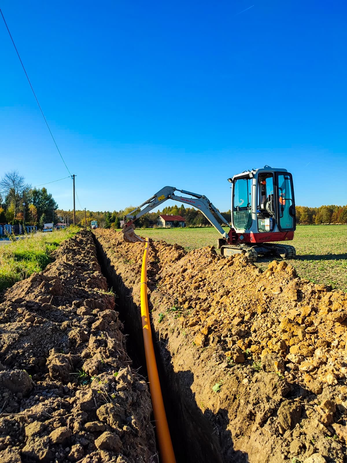 Żółty rurociąg ułożony w wykopie ziemnym, z małą koparką Takeuchi w tle na tle błękitnego nieba i zielonego pola.