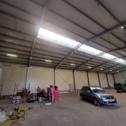 IHOR IVATSKO roboty elektryczne - Wnętrze hali z widoczną konstrukcją stalową dachu, oświetleniem, stertą materiałów izolacyjnych, stosem paczek Rockwool, zaparkowanym samochodem Suzuki SX4 i pracującym mężczyzną w tle.