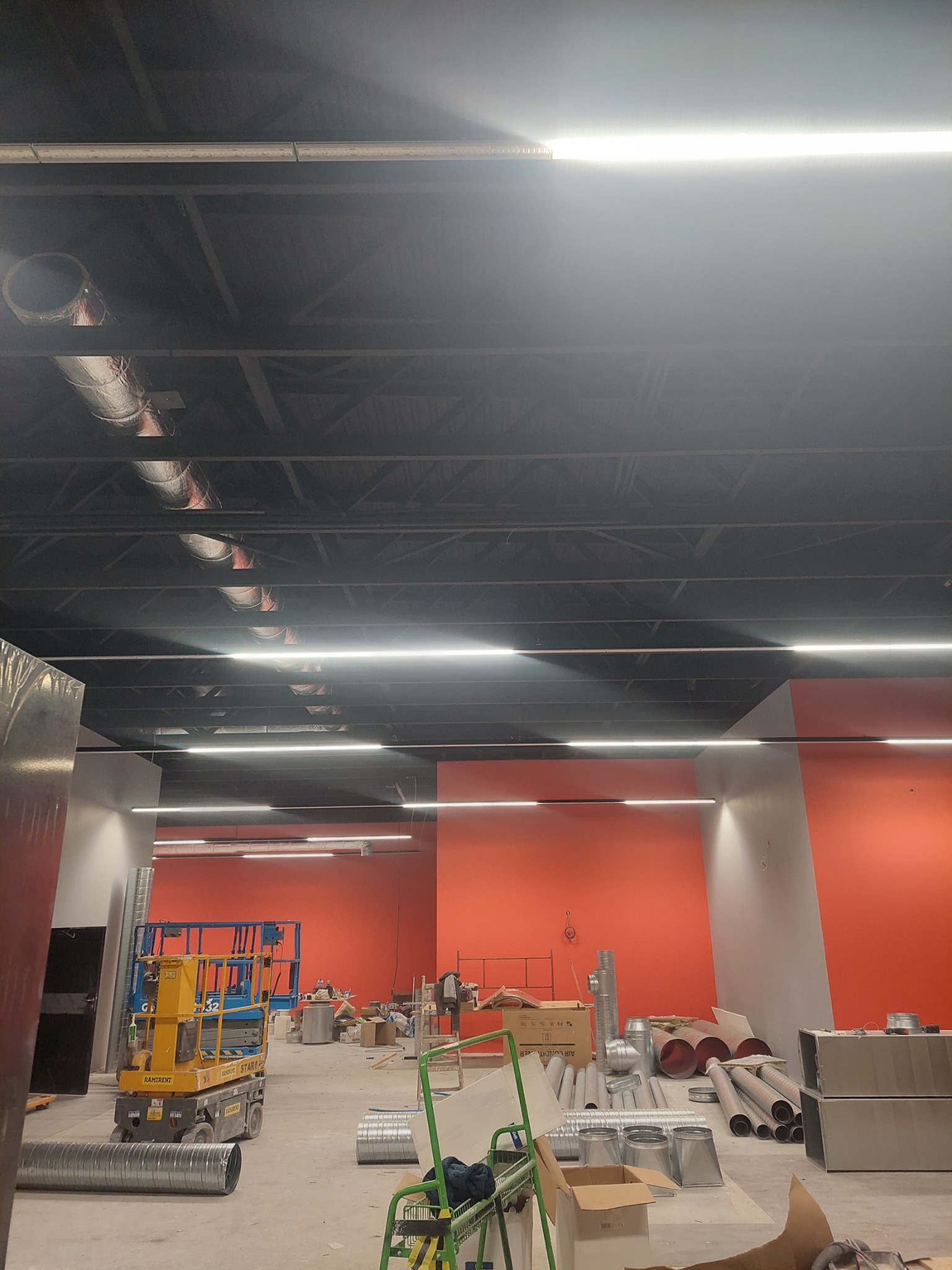 Wnętrze hali w trakcie instalacji wentylacji: widoczne rury wentylacyjne na podłodze i pod sufitem, podnośnik nożycowy, pomarańczowe ściany i oświetlenie LED.
