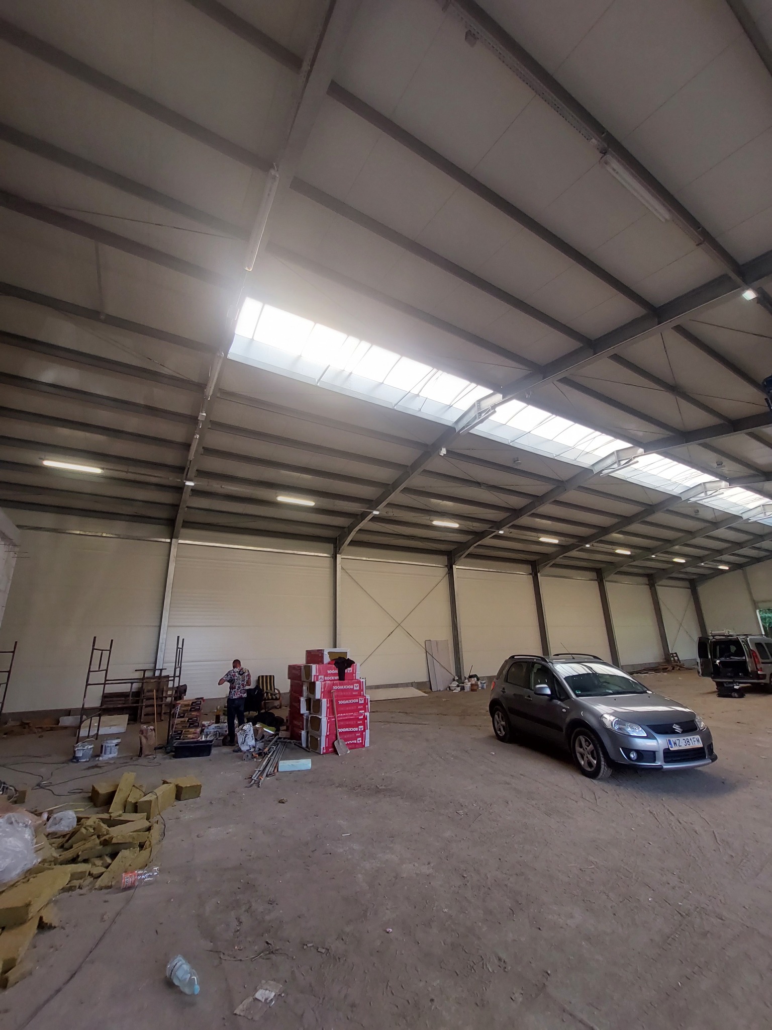 Wnętrze hali z widoczną konstrukcją stalową dachu, oświetleniem, stertą materiałów izolacyjnych, stosem paczek Rockwool, zaparkowanym samochodem Suzuki SX4 i pracującym mężczyzną w tle.