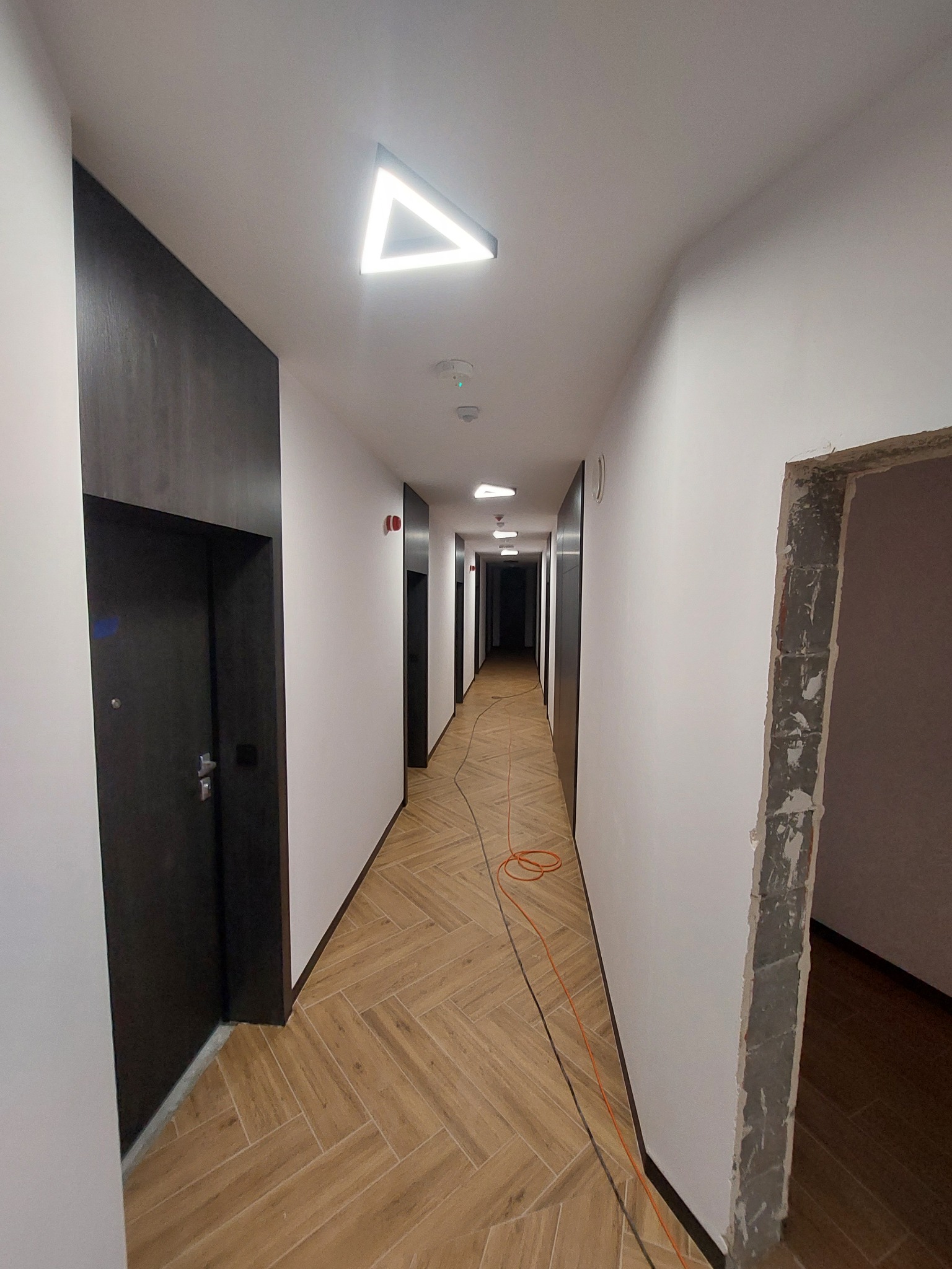 Długi korytarz w budynku mieszkalnym z drewnianą podłogą ułożoną w jodełkę, białymi ścianami, czarnymi drzwiami i trójkątną lampą sufitową; widoczne przewody elektryczne sugerują trwające prace...