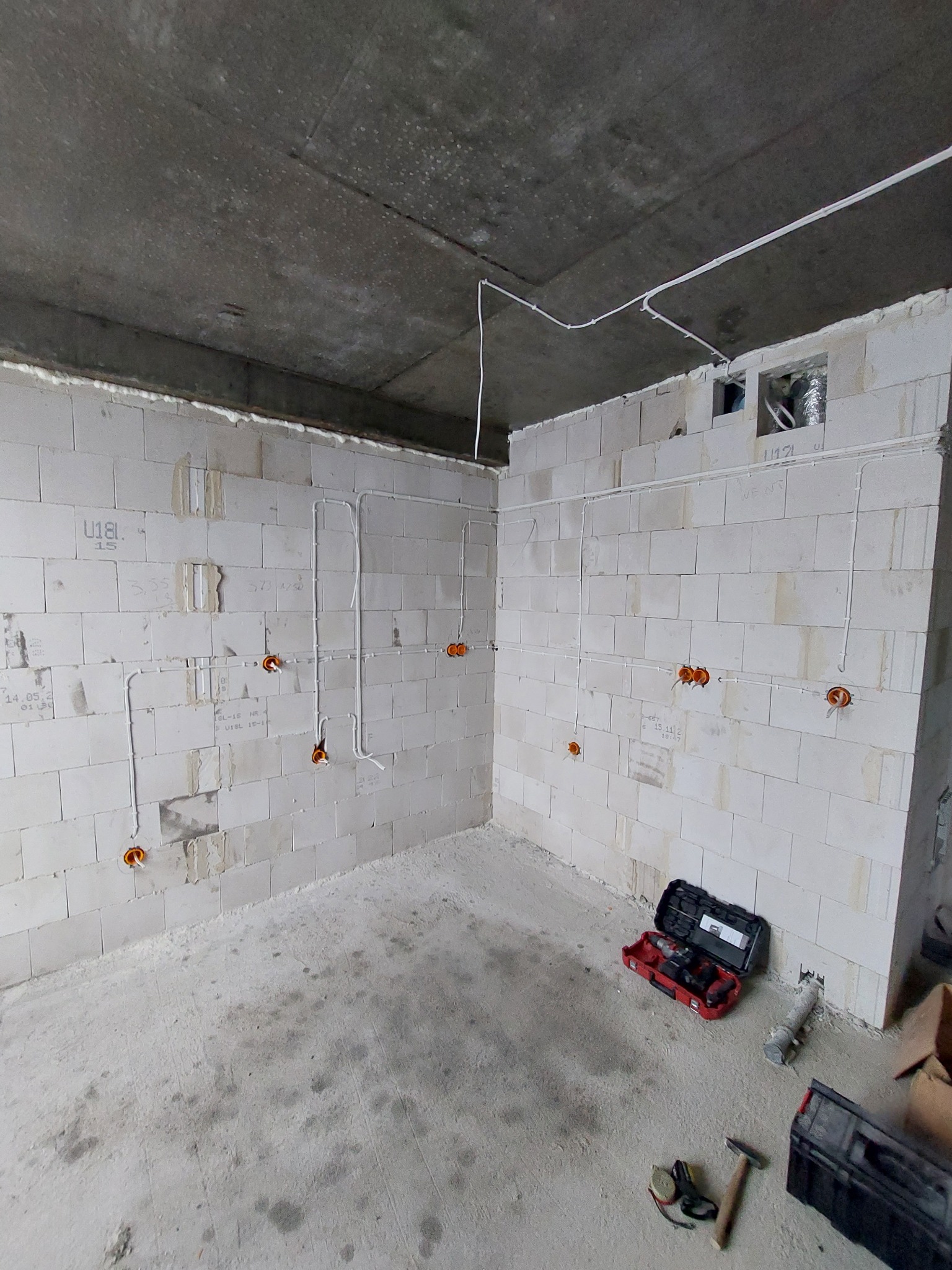 Surowy stan: ściany z betonu komórkowego z rozprowadzonymi przewodami elektrycznymi, puszki instalacyjne w kolorze pomarańczowym, kable prowadzone pod sufitem, skrzynki z narzędziami na podłodze.
