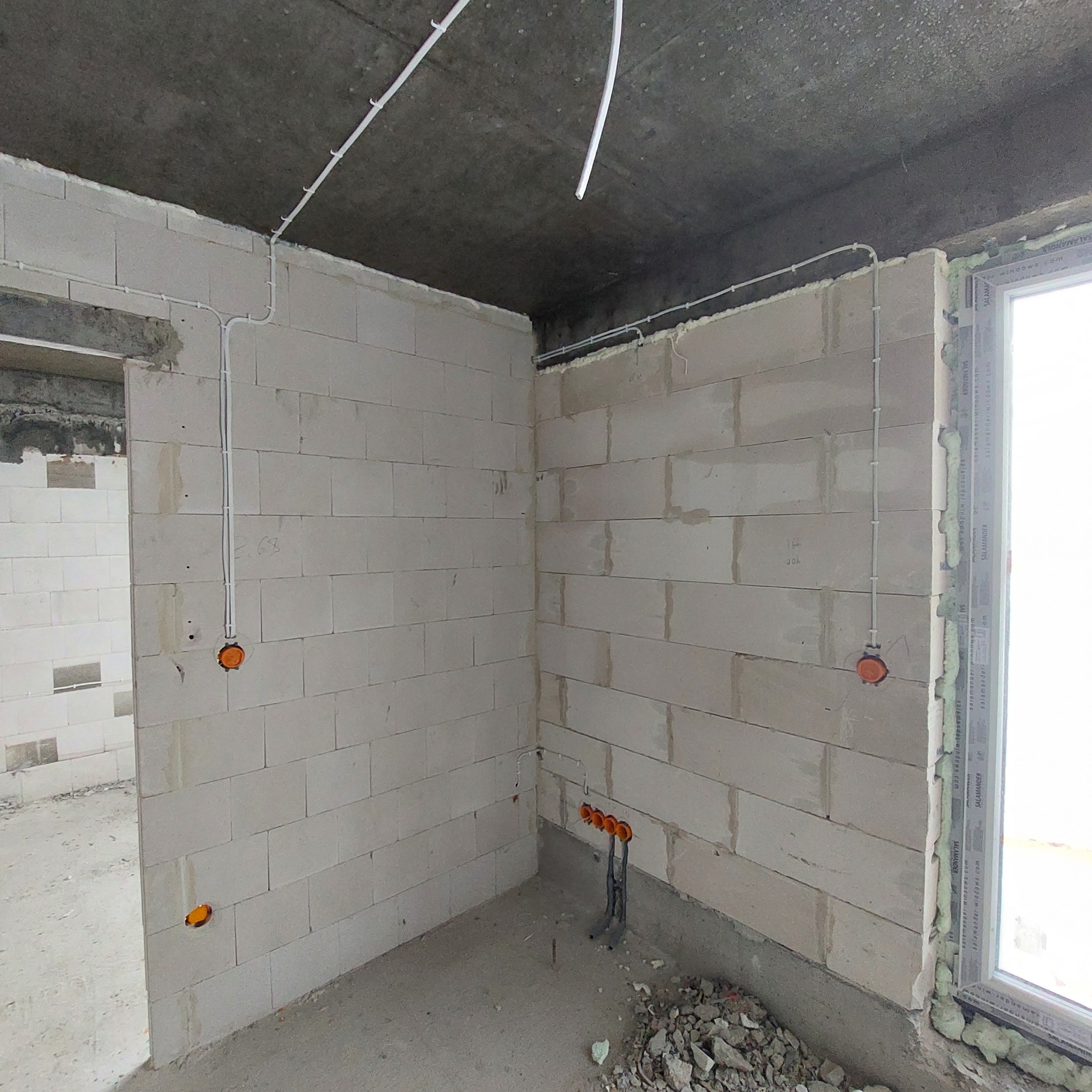 Surowy stan: Wnętrze budynku w trakcie prac, widoczne białe ściany z betonu komórkowego, wystające kable elektryczne w białych rurkach mocowanych do sufitu i ścian, puszki instalacyjne w kolorze...