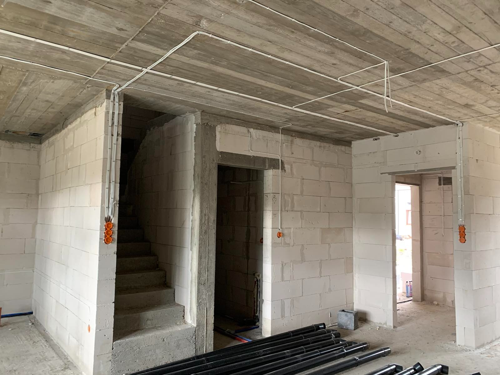 Surowy stan wnętrza budynku w trakcie rozprowadzania przewodów elektrycznych w peszlach, widoczne puszki instalacyjne i rury na podłodze.