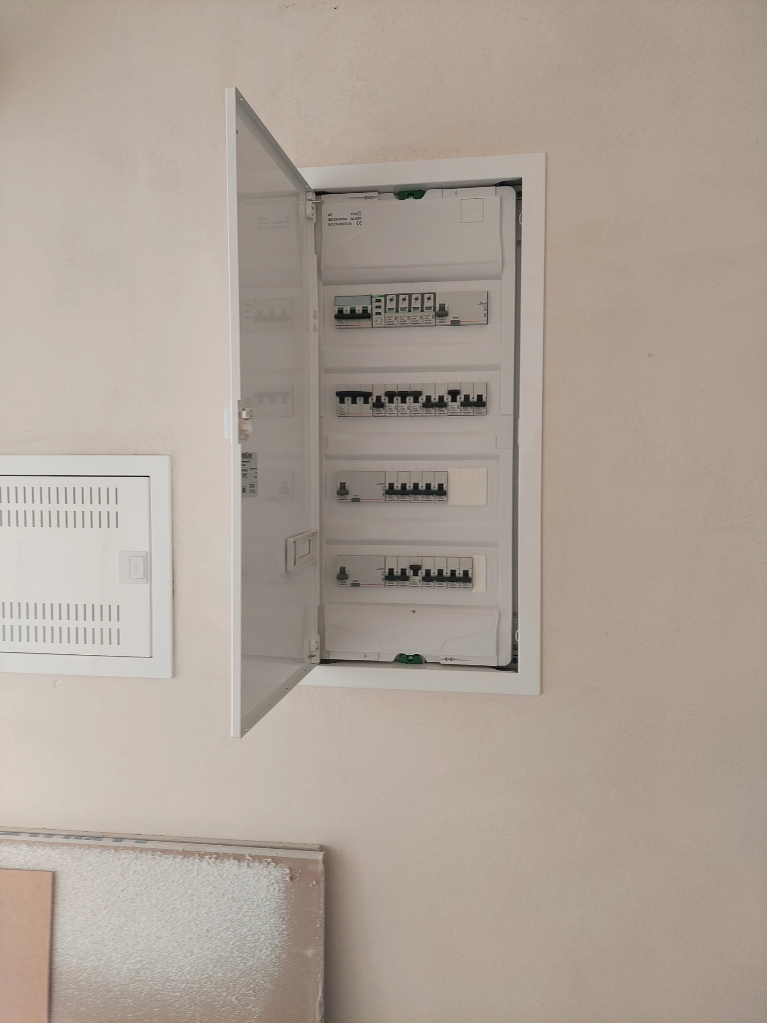 Otwarta, biała skrzynka z bezpiecznikami zamontowana na beżowej ścianie, widoczne rzędy wyłączników i etykiety, obok mniejsza skrzynka z kratką wentylacyjną.