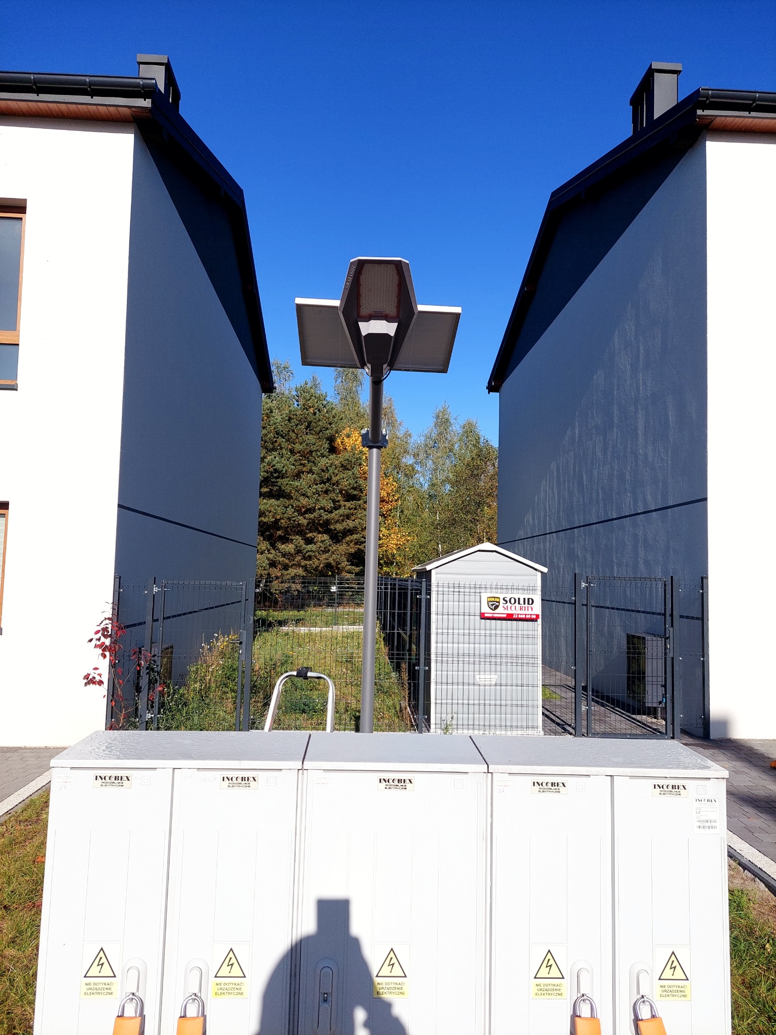 Zewnętrzna szafa elektryczna Inco Bex z ostrzeżeniami, na tle lampy solarnej z panelem fotowoltaicznym, budynku i budki Solid Security