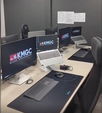 Stanowiska pracy z monitorami i laptopami z logo firmy na biurkach w biurze. Widoczne klawiatury, myszki i podkładki. Przygotowane do pracy.