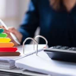 Sprawdź Dom - Kobieta w biurze oblicza wskaźniki energetyczne domu, używając kalkulatora i przeglądając dokumenty, z dekoracyjnym symbolem domu z ocenami energetycznymi A-G.