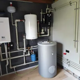 Kompletna instalacja grzewcza z kotłem Kaisai, zbiornikami i rozdzielaczami, rury prowadzone wzdłuż ścian. Nowoczesny system grzewczy w kotłowni.