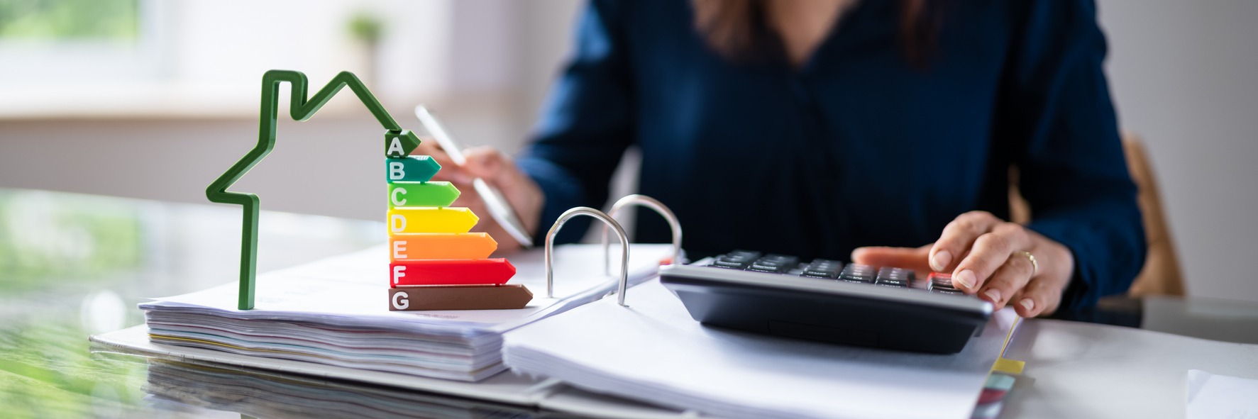 Kobieta w biurze oblicza wskaźniki energetyczne domu, używając kalkulatora i przeglądając dokumenty, z dekoracyjnym symbolem domu z ocenami energetycznymi A-G.