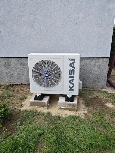 Kompaktowa, biała jednostka zewnętrzna klimatyzacji Kaisai, ustawiona na betonowych bloczkach, na tle szarej ściany budynku i trawnika.