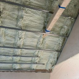 GREENFLOW SOLUTIONS SPÓŁKA Z OGRANICZONĄ ODPOWIEDZIALNOŚCIĄ - Świeżo nałożona, jasna pianka izolacyjna między metalowymi profilami sufitowymi, zabezpieczona folią malarską na ścianie, widoczny fragment okna.