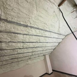 GREENFLOW SOLUTIONS SPÓŁKA Z OGRANICZONĄ ODPOWIEDZIALNOŚCIĄ - Poddasze po ociepleniu pianką PUR, widoczne metalowe elementy konstrukcyjne wystające z piany, ściana boczna pomalowana na jasny kolor, na podłodze resztki materiałów budowlanych w czarnym worku.