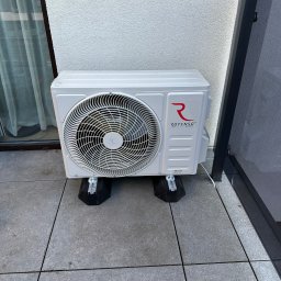 GREENFLOW SOLUTIONS SPÓŁKA Z OGRANICZONĄ ODPOWIEDZIALNOŚCIĄ - Zewnętrzna jednostka klimatyzacji Rotenso Teta Series, umieszczona na czarnych podkładkach antywibracyjnych na szarym tarasie, z widoczną ścianą budynku i fragmentem balustrady.