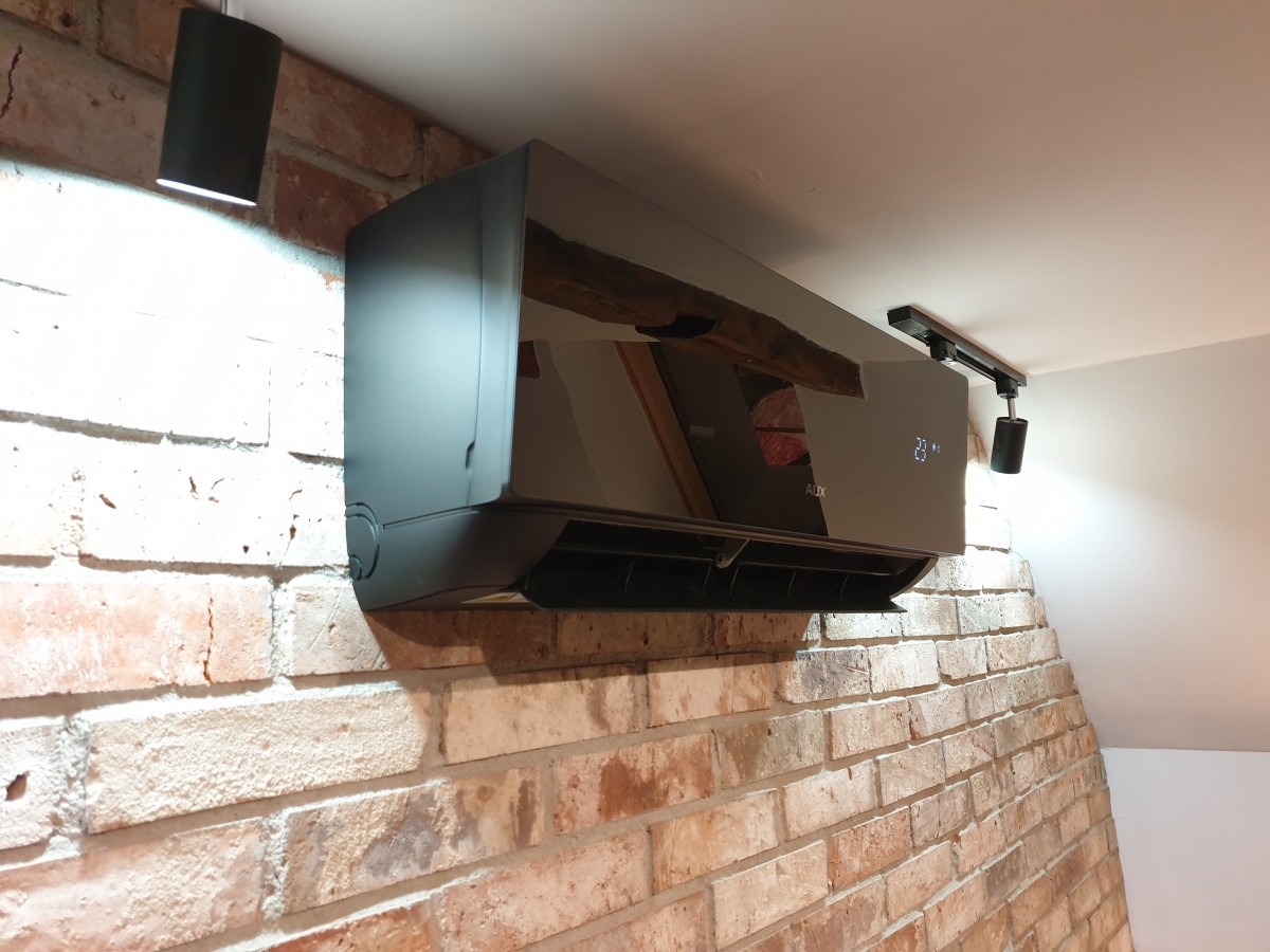 Czarny klimatyzator marki AUX zamontowany na ścianie z cegły, oświetlony dwoma reflektorami punktowymi.