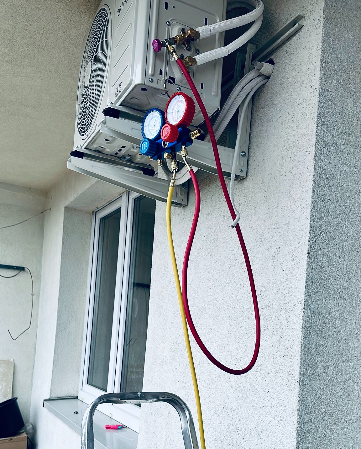 Biały klimatyzator zewnętrzny zamontowany na elewacji budynku, podłączony do manometrów serwisowych z czerwonymi i żółtymi przewodami, widoczna drabina i okno.