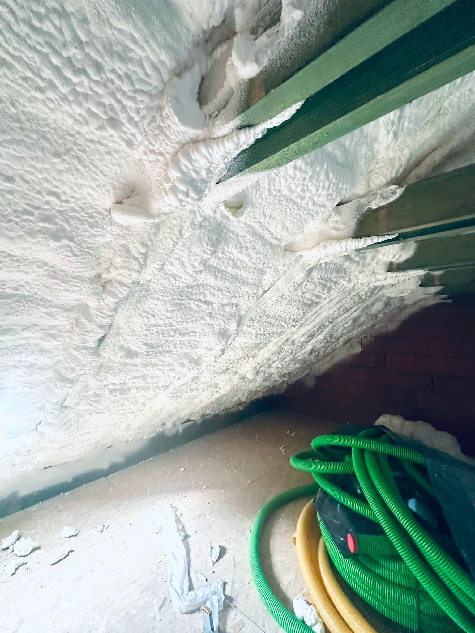 Poddasze ocieplone białą pianką poliuretanową, widoczne zielone belki konstrukcyjne i zwój zielonych węży na podłodze.